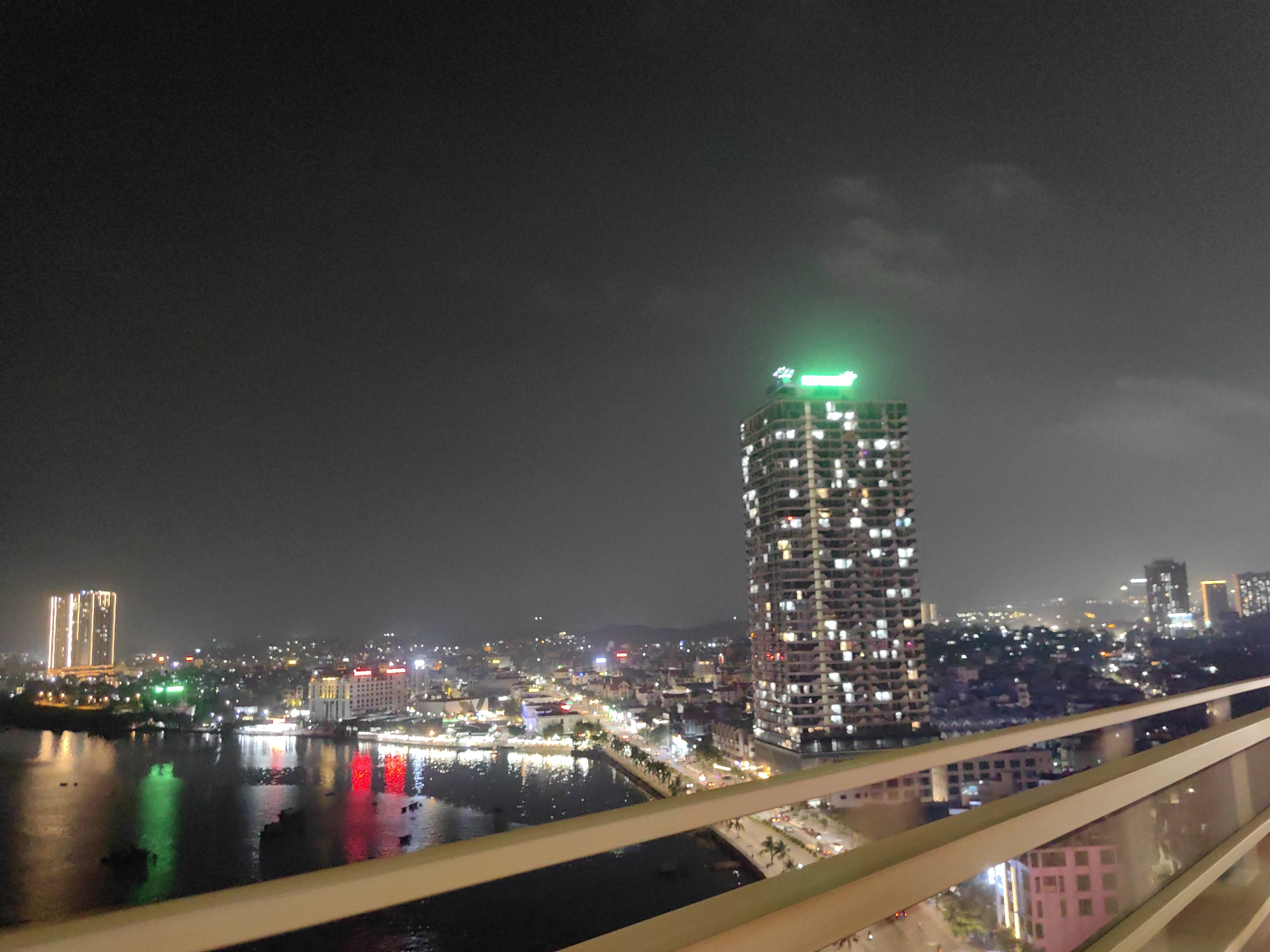 The image shows a nighttime cityscape in Hong Gai Ward, Vietnam. A tall, multi-story building with illuminated windows and a green light on top dominates the center-right of the frame. To the left, across a body of water, are other buildings, some with bright lights reflecting on the water's surface. A bridge spans the water, connecting different parts of the city. In the foreground, a railing is visible, suggesting the photo was taken from a high vantage point. The sky is dark, with some subtle cloud formations. The overall scene is lit by artificial lights, highlighting the urban environment.