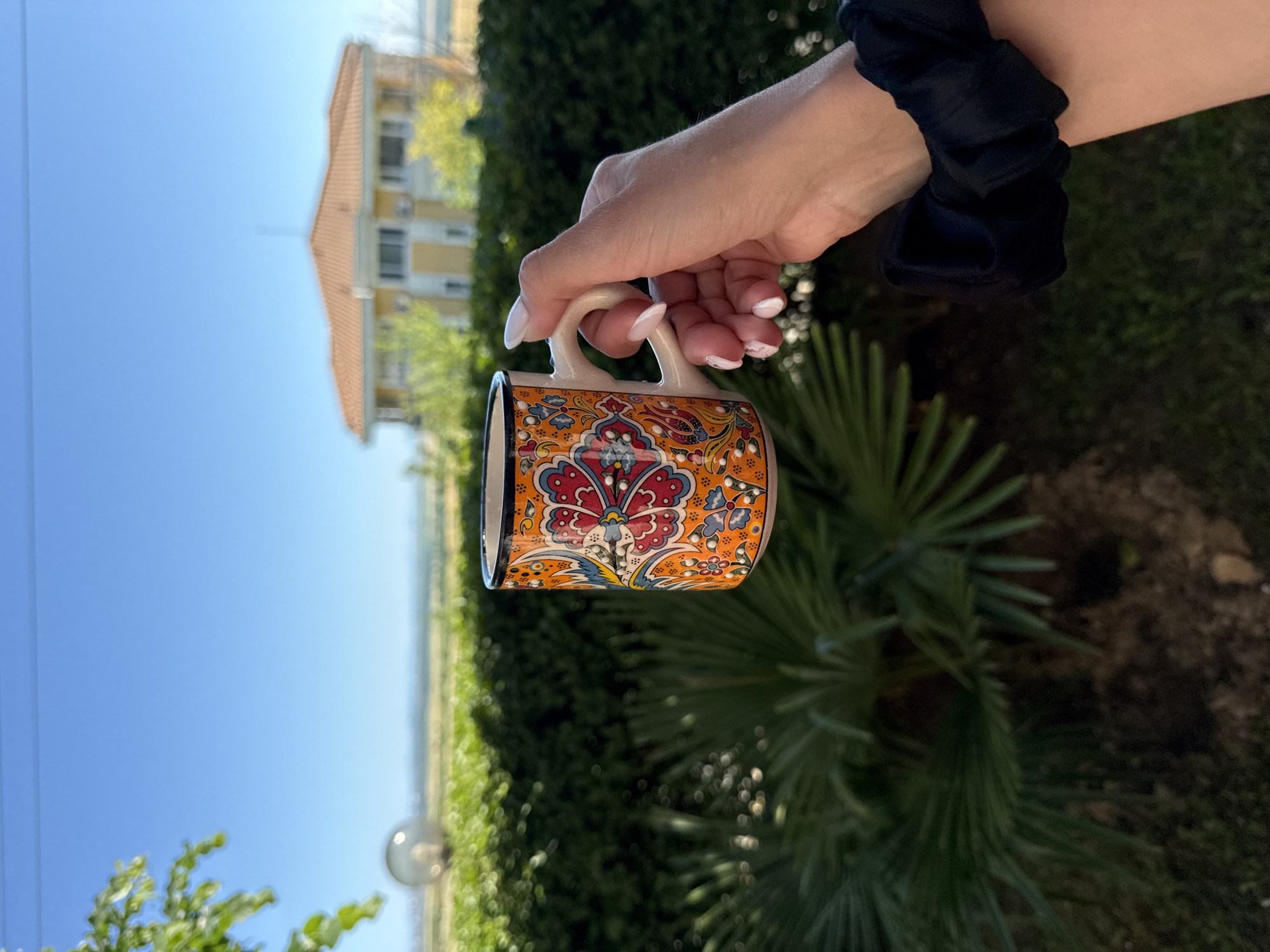 In Kosharitsa, Bulgaria, a hand with manicured nails is holding a decorative mug. The mug is white with an orange base and adorned with colorful floral patterns in red, blue, and yellow. The background features a clear blue sky above a manicured green hedge and palm fronds. In the distance, a yellow and orange tiled-roof house is visible. The overall scene appears to be set outdoors in a sunny, tranquil environment.