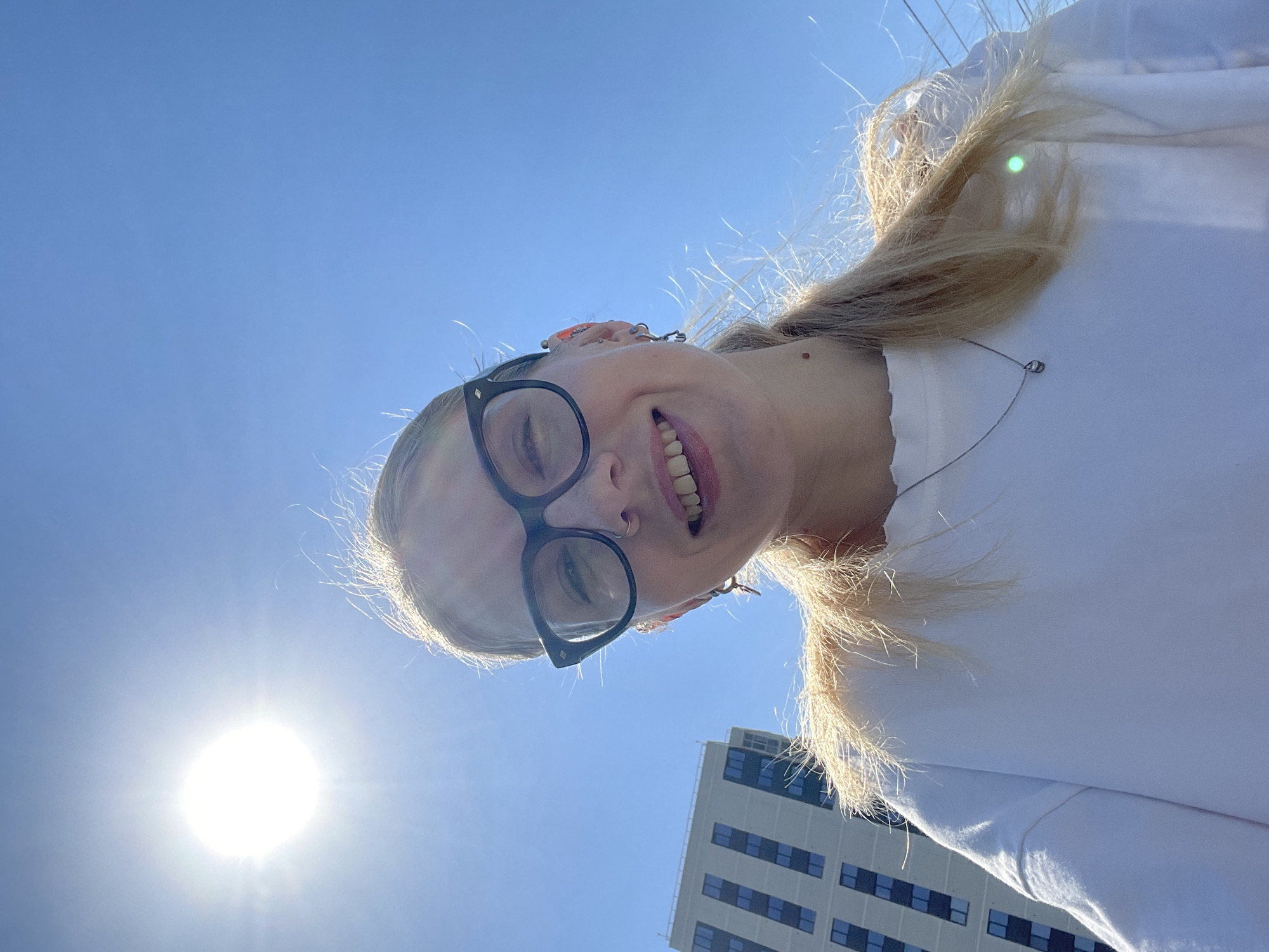 A low-angle shot looking up at a woman with blonde hair, wearing black glasses and a white shirt, against a clear blue sky with the sun visible in the lower-left corner. Part of a light-colored building with numerous dark windows can be seen in the lower-right. The location is Izhevsk, Russia, as evidenced by the building's architecture and style, typical of urban areas in that region. The woman is smiling and looking down towards the camera.