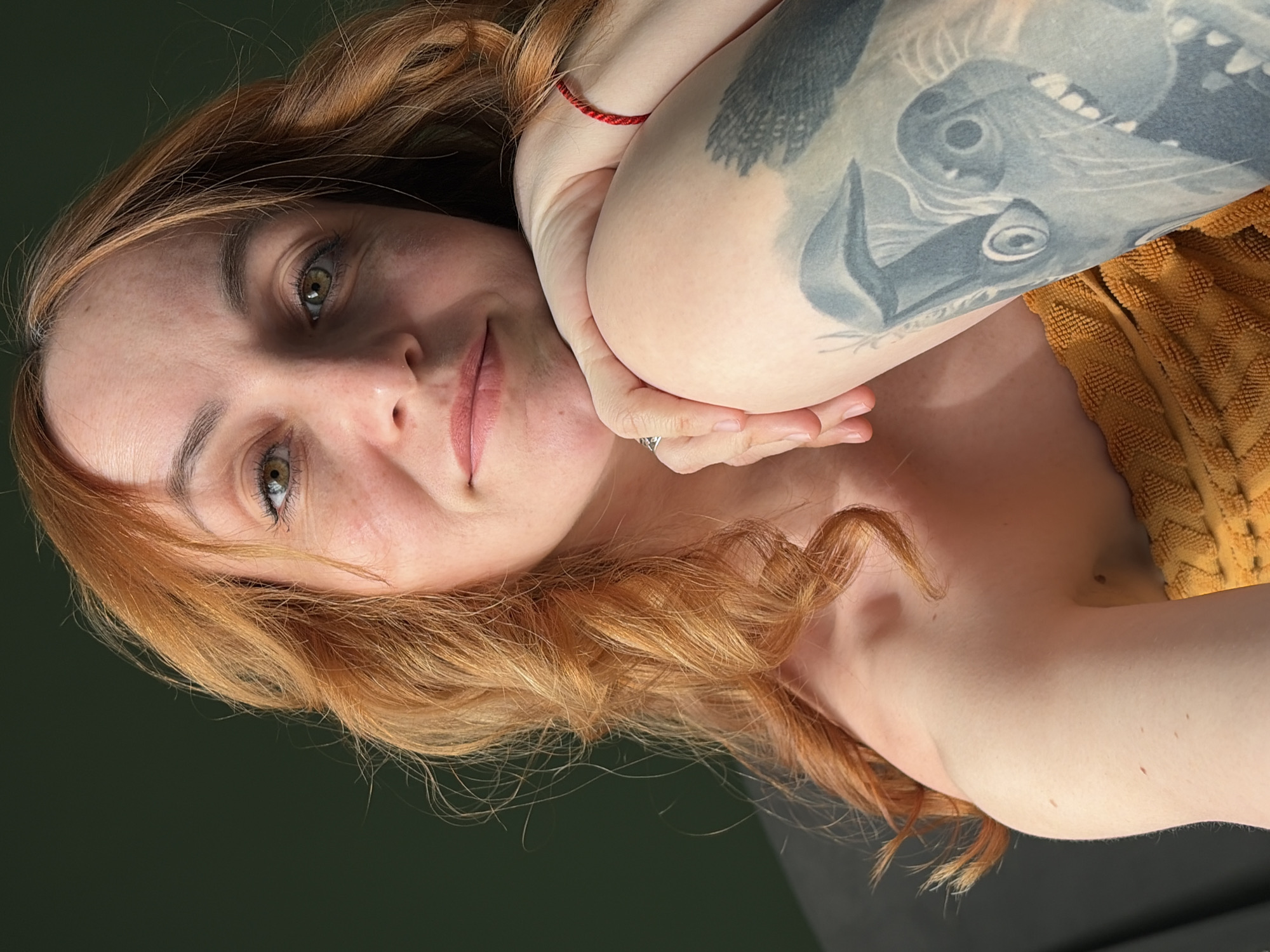 A woman with red hair and light skin is looking at the camera. She has green eyes and is lightly smiling. Her right arm is bent, and her elbow is touching her face. A red string is visible on her wrist. There is a tattoo of a bird on her upper arm. She is wearing a yellow textured top. The background is dark and out of focus. There is no visible information to determine the location in the city of Chaikovsky, Russia.