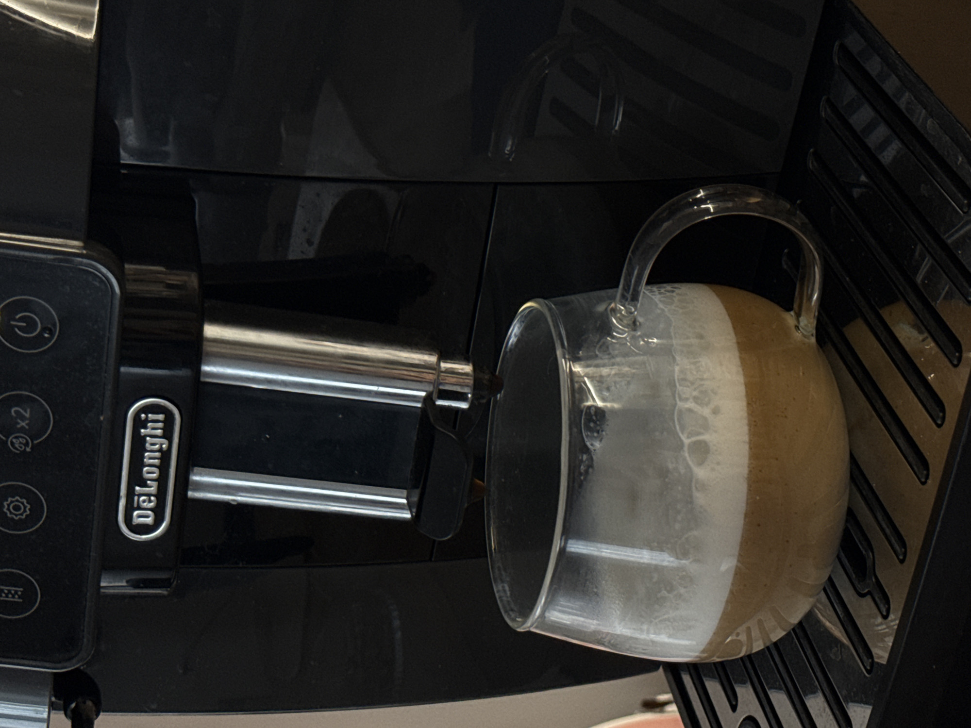 A close-up, slightly angled, high-contrast photograph depicts a Delonghi automatic espresso machine dispensing espresso and milk foam into a glass mug with a clear handle. The machine is primarily black. The mug sits on a ribbed, black plastic drip tray. The location is not determinable from the image.