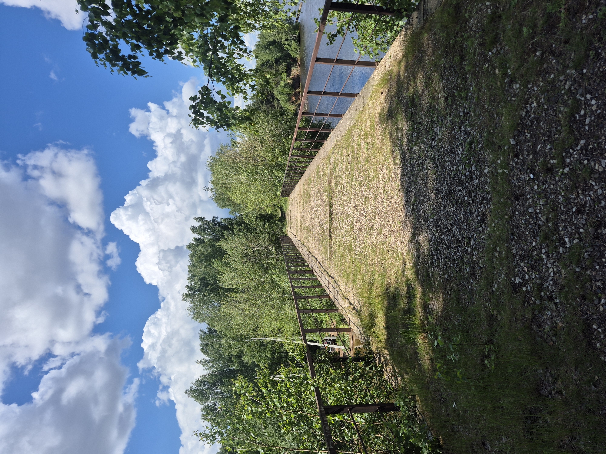 The image shows an elevated pathway with a metal railing, possibly a former railway embankment, located in Chaikovsky, Russia. The pathway is covered in gravel and runs along a river, visible through the railing. Lush green trees and vegetation line the pathway on one side. On the other side, the ground slopes down, covered in grass and gravel. The sky is bright blue with large, puffy white clouds. The image appears to be taken during the day, with strong sunlight casting shadows on the pathway.