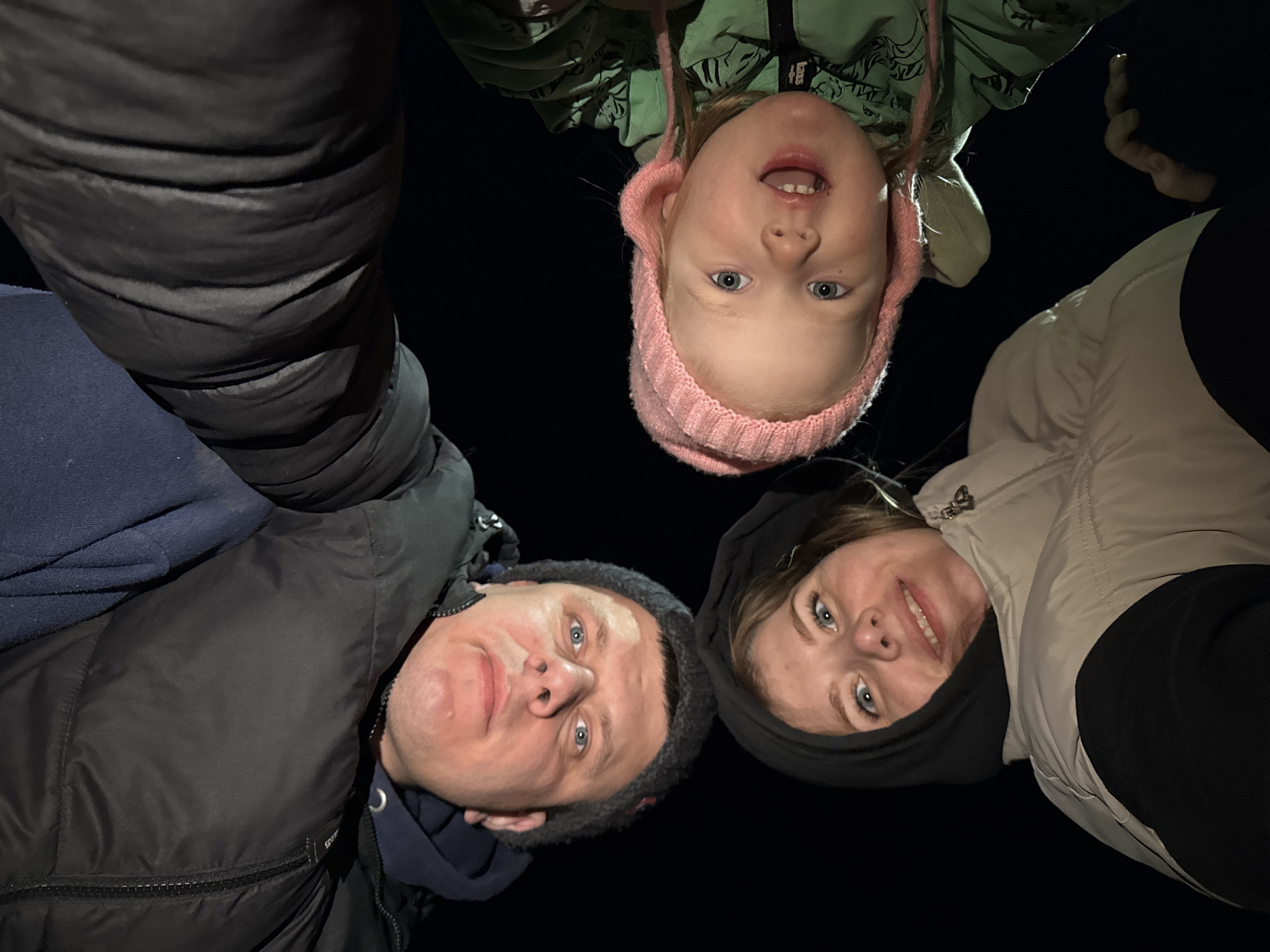 The image shows four people in Gornyi, Dvurechenskoe S/P, Russia. From left to right: a man wearing a black padded jacket over a blue garment and a grey hat, a woman in a beige padded jacket over a black hoodie, a young girl in a pink hat and a green jacket. All are looking directly at the camera. The background is completely dark, indicating an outdoor setting at night. They appear to be huddled together.