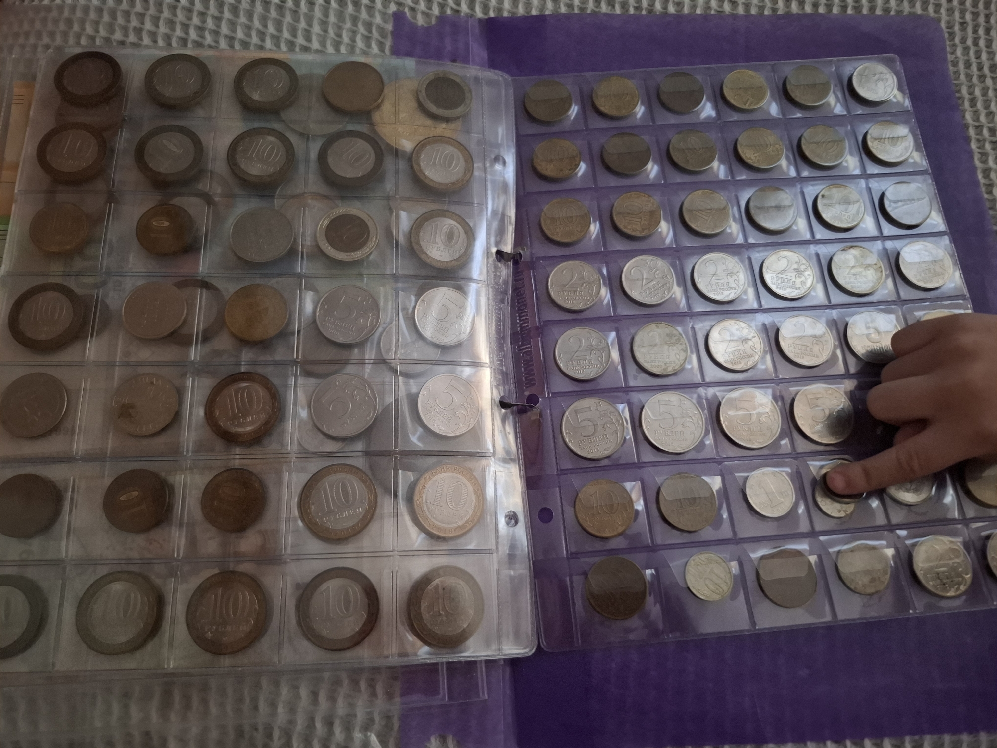 The image shows two coin collection pages from a coin collecting album. The coin collection pages are partially visible, and the album is on a gray and purple surface. The left page has a clear plastic cover, while the right page is a purple plastic. The coins in the pages are different colors and denominations, some are inscribed with "10 рублей". A person's hand is visible on the right, touching a coin on the right page. The coins on the right page have different inscriptions and dates. The image shows a collection of Russian coins and a person handling them. It is not possible to determine the exact location within Amzya, Russia, from the image alone.