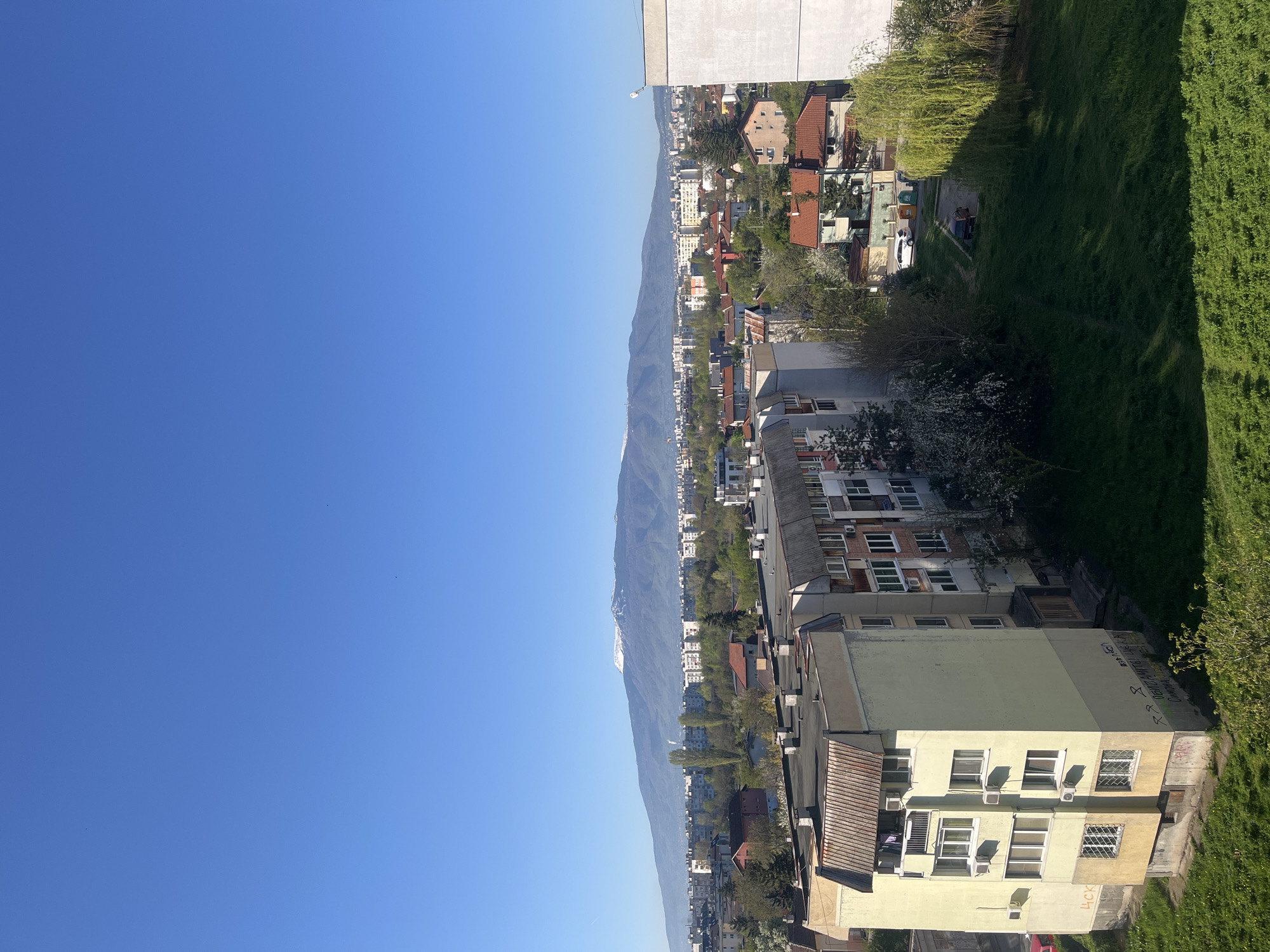 The image shows a cityscape in Sofia, Bulgaria, with a clear blue sky dominating the left half. The right half reveals an elevated view of residential buildings, trees, and grass. A mountain range is visible in the distance. The buildings are a mix of older and newer constructions with varying colors, including yellow and beige. There are signs of vegetation amongst the buildings, indicating a mix of urban and natural elements. The image is taken from a high vantage point, providing a broad overview of the urban landscape against the backdrop of the distant mountains. There are no people visible.