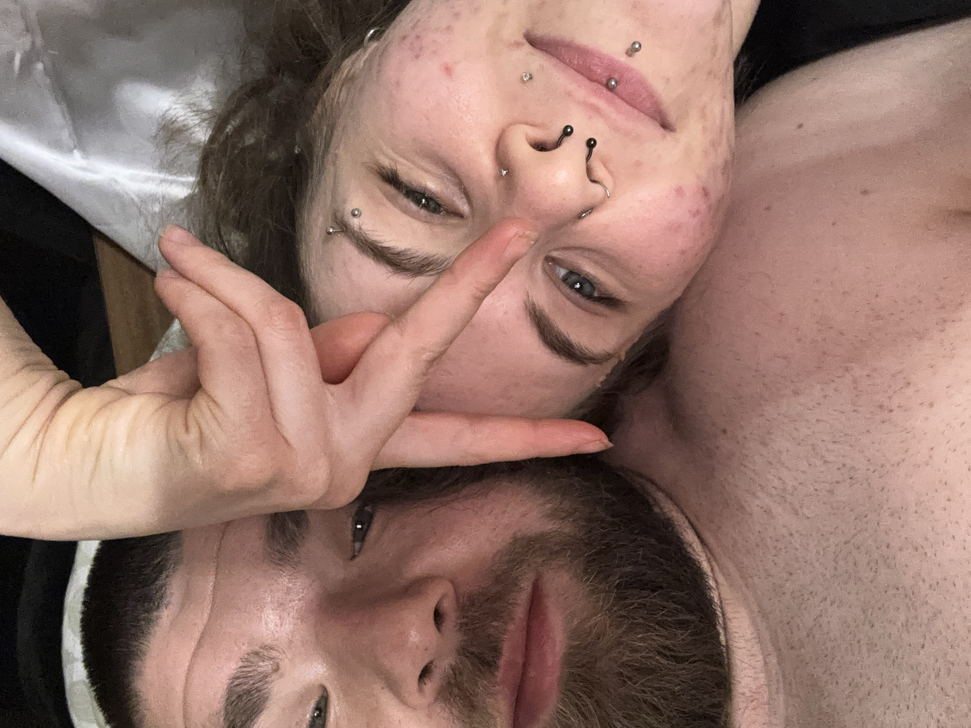The image shows two people lying down in close proximity, with the woman positioned above and to the left of the man. The woman has multiple facial piercings, including nose rings, eyebrow piercings, and lip piercings. She is making a peace sign with her hand, which is positioned over the man's face. The man has a beard and is lying with his head tilted back. The background appears to be a bed with white sheets. The specific location within the city of Fulda, Germany, cannot be determined from this image alone.