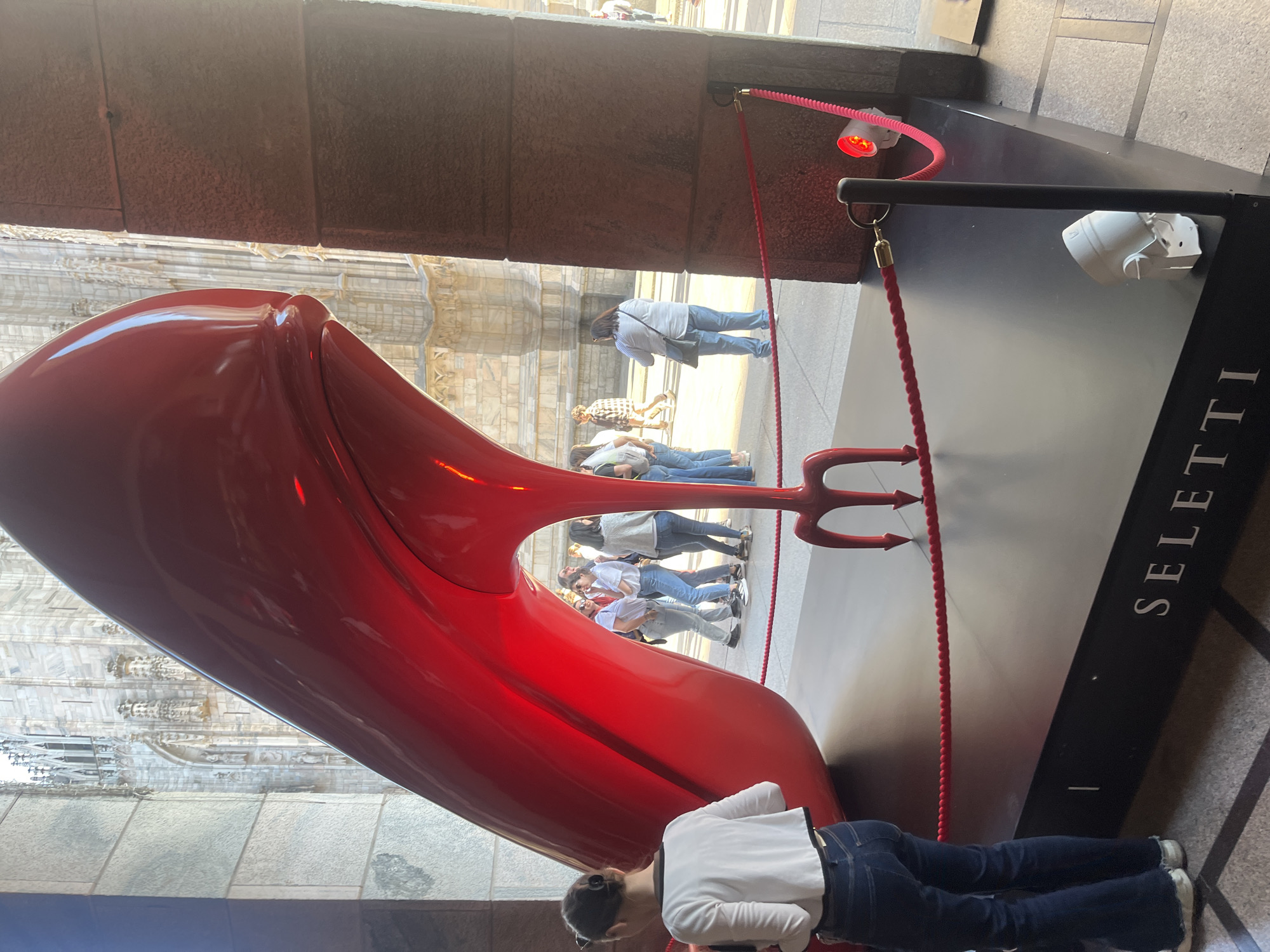 A large, glossy red sculpture is prominently displayed outdoors, likely as part of an art exhibition. The sculpture is an abstract, elongated form that evokes a high-heeled shoe or a stylized object, featuring a distinctive red trident shape at its base or heel. Its reflective surface distorts the surrounding environment.
In the foreground, a person wearing a white shirt and blue jeans is bending down, facing the sculpture. Behind the sculpture, several other individuals, mostly adults, are visible, either observing the artwork or passing by.
The sculpture rests on a black display platform, on which the word "SELETTI" is printed vertically in white capital letters. Red ropes with gold-colored stanchions are set up to delineate the display area. A small white speaker and a pink coiled cable with a red illuminated component are also present on the platform.
The background reveals a magnificent, ornate stone building, characterized by intricate carvings, columns, and spires, strongly suggesting a historical cathedral or a grand architectural landmark. The scene is brightly lit, indicating it is daytime. The overall impression is a public art installation situated in a busy, historically significant urban setting.
