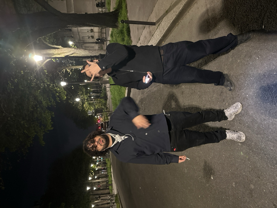 Two adult males are depicted standing on a paved, tree-lined street at night in an urban area identified as Paris, France, based on the characteristic Haussmannian architecture and streetlights.
The male on the left, with dark curly hair and a beard, wears a dark collared shirt, black pants, and patterned athletic shoes. He is adorned with several large, reflective necklaces, and a white earbud wire hangs from his neck. He holds a lit cigarette in his left hand, positioned at his side. His right arm is partially blurred, suggesting motion. He faces the camera directly with a neutral expression.
The male on the right, wearing dark-rimmed glasses, a dark zipped jacket, dark pants, and dark shoes, stands slightly angled to the camera. His right hand is raised, forming a gesture with extended index and pinky fingers. His left hand holds a small, illuminated object, consistent with a smartphone's flashlight, directed downwards.
The background features multiple vintage-style streetlights casting yellow-orange light along both sides of the street, which is lined with mature trees. Behind the trees on the right, light-colored, multi-story buildings with classic Parisian architectural elements are visible. Black metal bollards are present along the curb on the right side of the street.