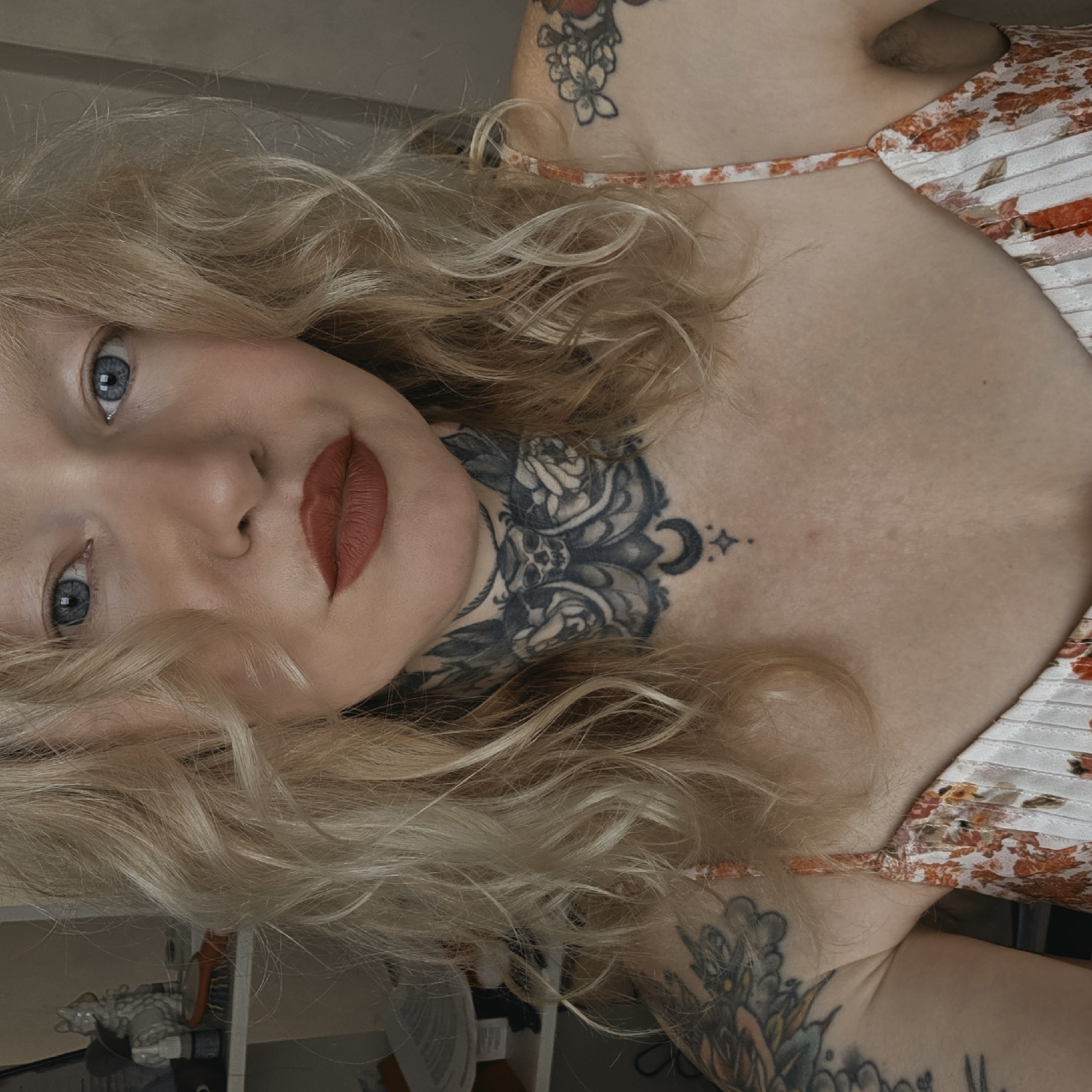 A woman with fair skin, light blonde curly hair, and blue eyes is posing indoors. She has red lipstick on. A large black tattoo covers her neck and upper chest. She is wearing a floral-patterned, light-colored dress with thin straps. She has tattoos on her arms as well. The background shows indistinct objects in a cluttered space. There's no indication of a location in St. Petersburg, Russia.