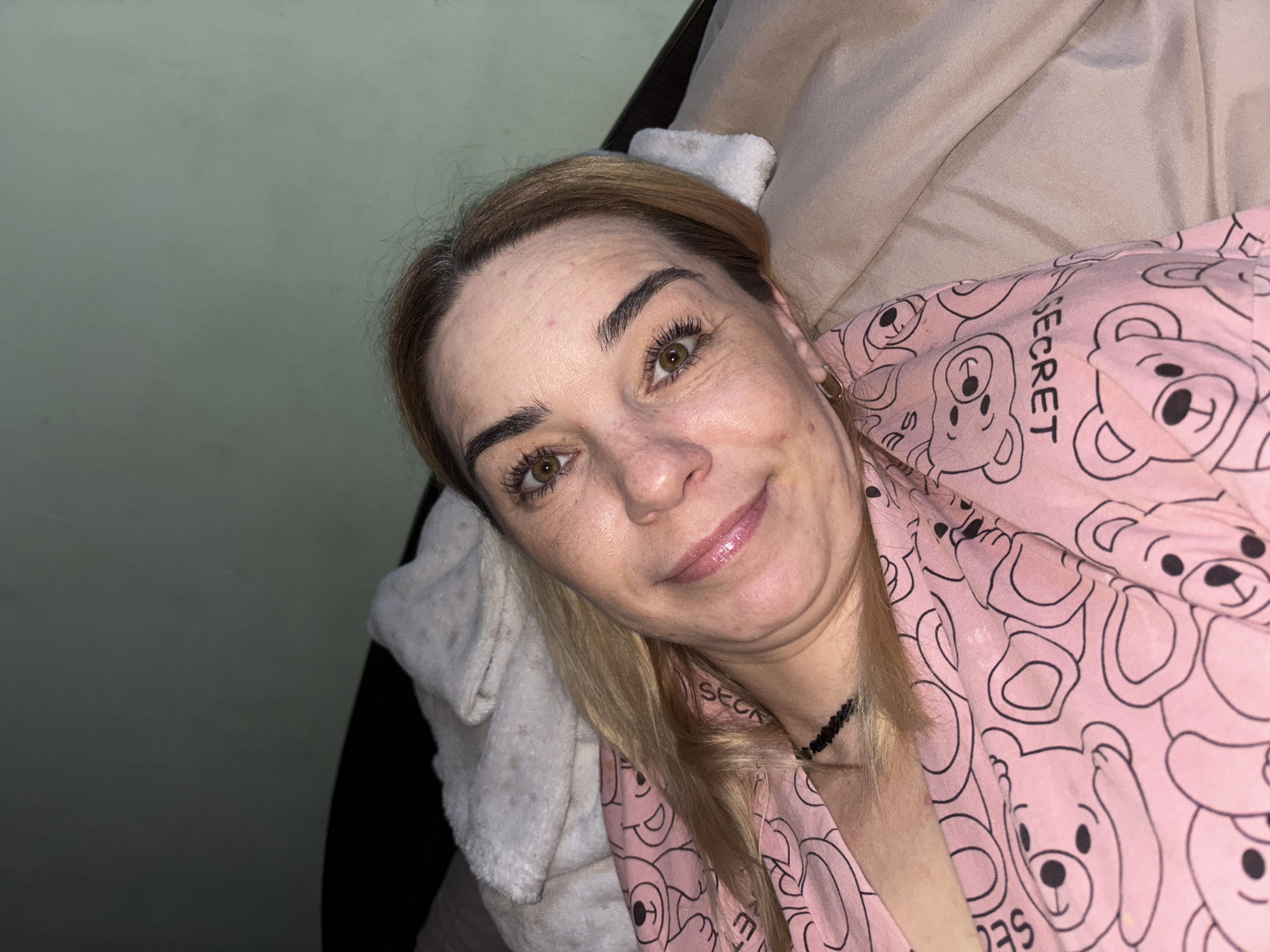 The image shows a close-up, waist-up, selfie-style photograph of a blonde woman lying down. She is wearing pink pajamas adorned with a teddy bear print and the word "SECRET." A dark, beaded necklace is visible around her neck. Her head is tilted, and she is looking directly at the camera with a slight smile. The background consists of a light green wall and bedding in neutral tones, including a folded white blanket. The lighting suggests an indoor setting, possibly a bedroom, with no indication of an outdoor or urban environment. The location of Unknown, Russia is not ascertainable from the visual information provided.