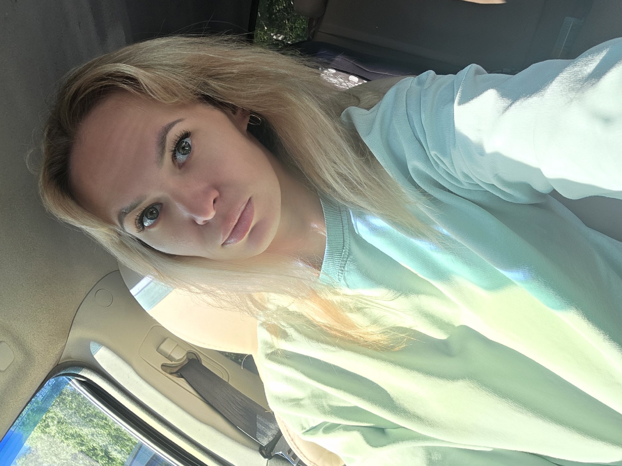 A blonde woman is captured in a selfie taken from inside a car. She is wearing a light teal sweatshirt. The sunlight streams in from the left, illuminating the interior of the car and casting shadows across her face and clothing. The background shows the car's interior with a seatbelt visible. The scene is set outdoors, with greenery visible through the car window, suggesting a location within the city of Chaikovsky, Russia.