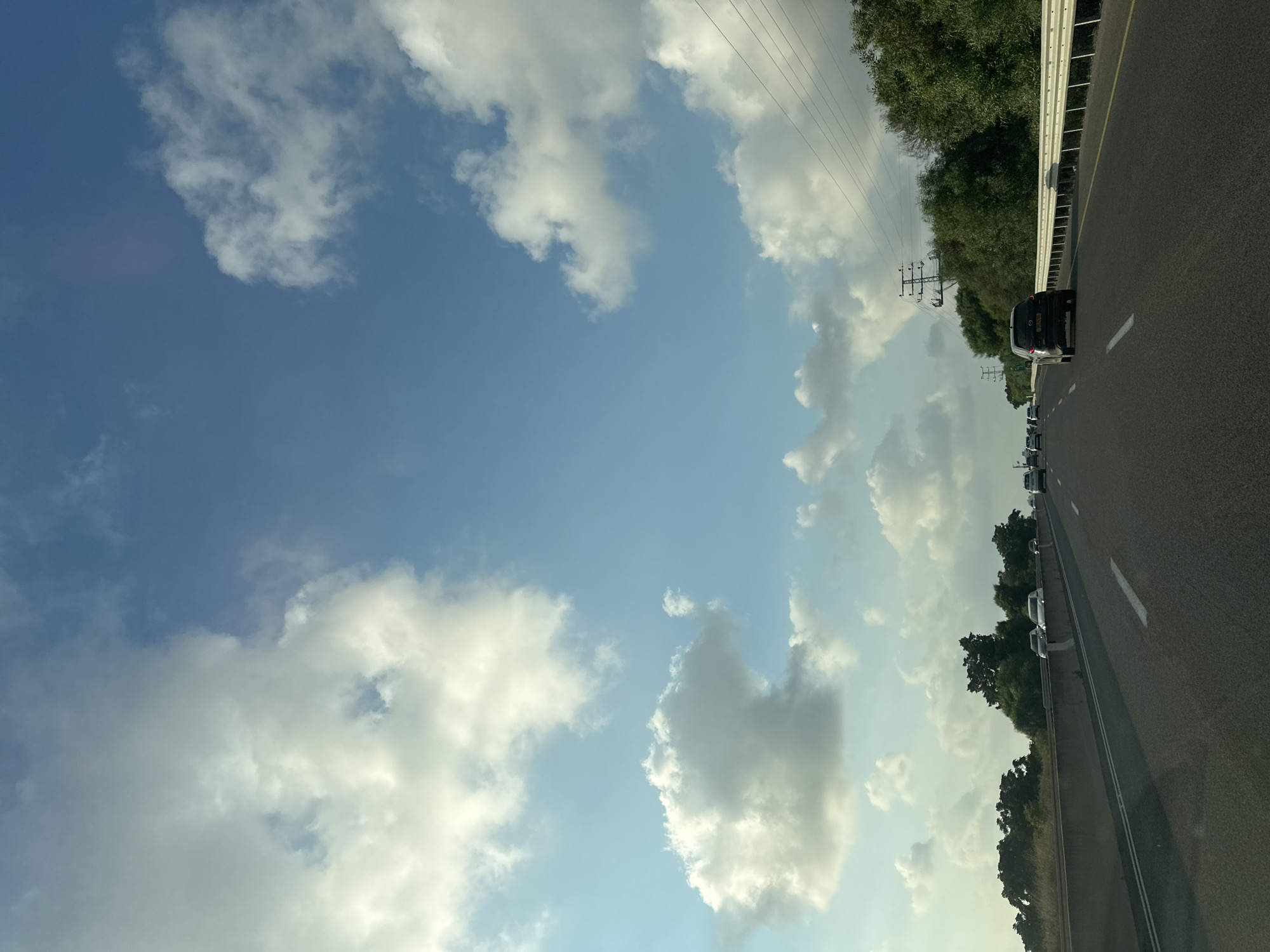 The image captures a perspective from a moving vehicle on a multi-lane highway in the Mate Asher Regional Council, Israel. The sky is a vibrant blue, dotted with large, fluffy white clouds. On the right side of the highway, a dense line of green trees borders the road, with power lines and utility poles visible above them. On the left, a concrete barrier separates the highway from an area with dry grass and more trees. Several vehicles, including cars and a dark SUV in the foreground, are traveling in the same direction. The road has clear white lane markings. The scene suggests daytime travel under a partly cloudy sky.