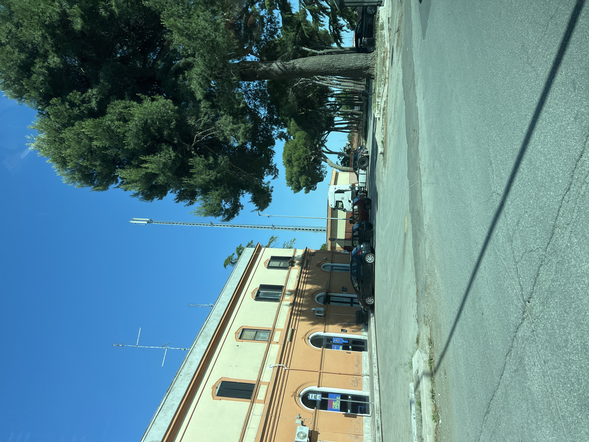 The image displays an outdoor scene in Santeramo in Colle, Italy, captured on a clear, sunny day under a bright blue sky, possibly from within a vehicle. On the left, a two-story building features a light yellow upper facade with four dark-shuttered windows and an orange-brown brick-textured lower facade with multiple arched entrances. Above the leftmost entrance, an air conditioning unit is mounted, alongside a blue bus stop sign and a vertical sign partially legible as "IDEZ IN MOVIMENT" above "ATM." Another air conditioning unit is present further right on the lower facade. The building's roofline is adorned with multiple antenna structures, including a prominent multi-element antenna.
In the midground, a paved area serves as a parking lot, where a black compact car is parked directly in front of the building. To its right, a dark-colored older model car is partially visible. Further right and slightly behind, a white semi-truck tractor unit with a dark chassis is parked. On the far right, another dark sedan is parked partially obscured by a large, mature pine tree that dominates the right side of the frame with its thick trunk and dense green canopy. A line of similar pine trees extends into the background, providing shade. Behind these trees, a tall cellular tower with multiple antennas is distinctly visible.
The foreground consists of an asphalt road showing several cracks and wear, separated from the parking area by a low concrete curb or planter. A long, thin shadow, likely from an unseen utility pole or antenna mast, stretches diagonally across the road from the upper left to the lower right. On the far left, a small, reddish-brown older car is partially visible behind the curb. No people or active interactions are visible in the scene.
