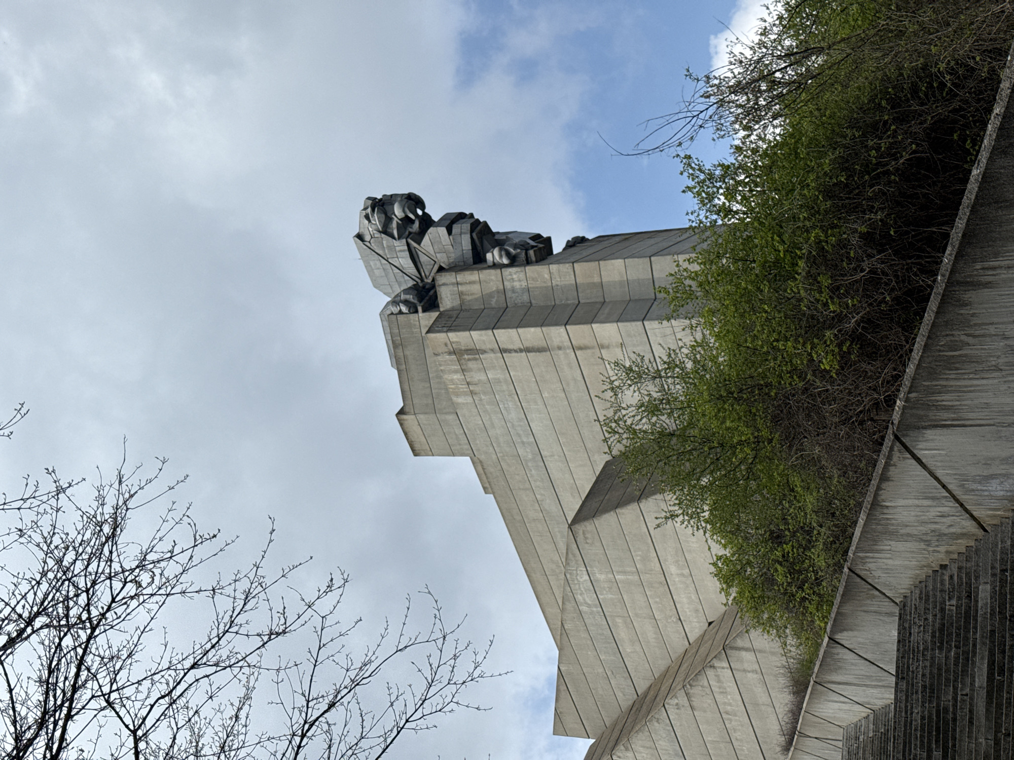 The image shows a low-angle view of the Monument to 1300 Years of Bulgaria, a large concrete structure in Shumen, Bulgaria. The monument features a massive stylized lion sculpture at its apex. The sky is partially cloudy with patches of blue. Vegetation, including trees and bushes, is visible framing the monument on both sides. A set of stairs lead up along the right side of the structure.