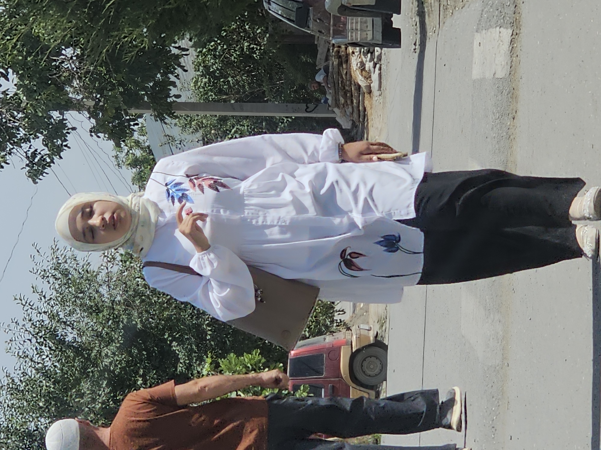 A middle-aged woman wearing a light-colored hijab and a white long-sleeved tunic with floral embellishments, paired with black pants and white shoes, walks along a paved street in Ögön, Kyrgyzstan. She carries a brown shoulder bag. To her left, a man in a brown t-shirt and dark pants walks away from the camera, wearing a white skullcap. In the background, a small red SUV is parked on the side of the road. The environment appears to be a residential or semi-urban area with trees and a utility pole visible.