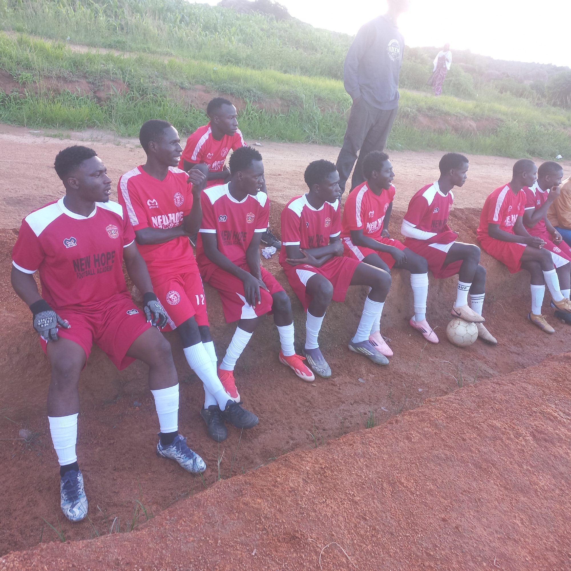 In Jos, Nigeria, a group of young men, likely a football team, are sitting on a raised dirt embankment. They are all wearing red and white football kits with the inscription "NEW HOPE FOOTBALL ACADEMY" visible on some of the shirts. The players are mostly young adults, with dark skin and short, dark hair. Some are looking towards the camera, while others are looking away. There is a football at the feet of one of the players. In the background, there is a grassy hillside and a person in the distance wearing a white and purple outfit. The lighting suggests it is late afternoon or early evening. The overall scene depicts a pause in a football activity, possibly a practice or a break during a match, in an outdoor setting characteristic of Jos.