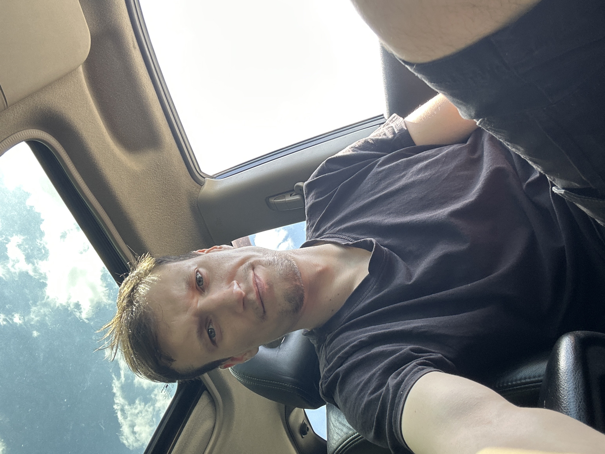In Makiv, Ukraine, a man sits in the passenger seat of a car, looking directly at the camera. He is wearing a black t-shirt and dark shorts. The car has a sunroof open, revealing a bright blue sky with white clouds. The interior of the car, including the dashboard and seats, is visible. The man has short, messy brown hair and a light beard. The overall lighting suggests it is daytime.