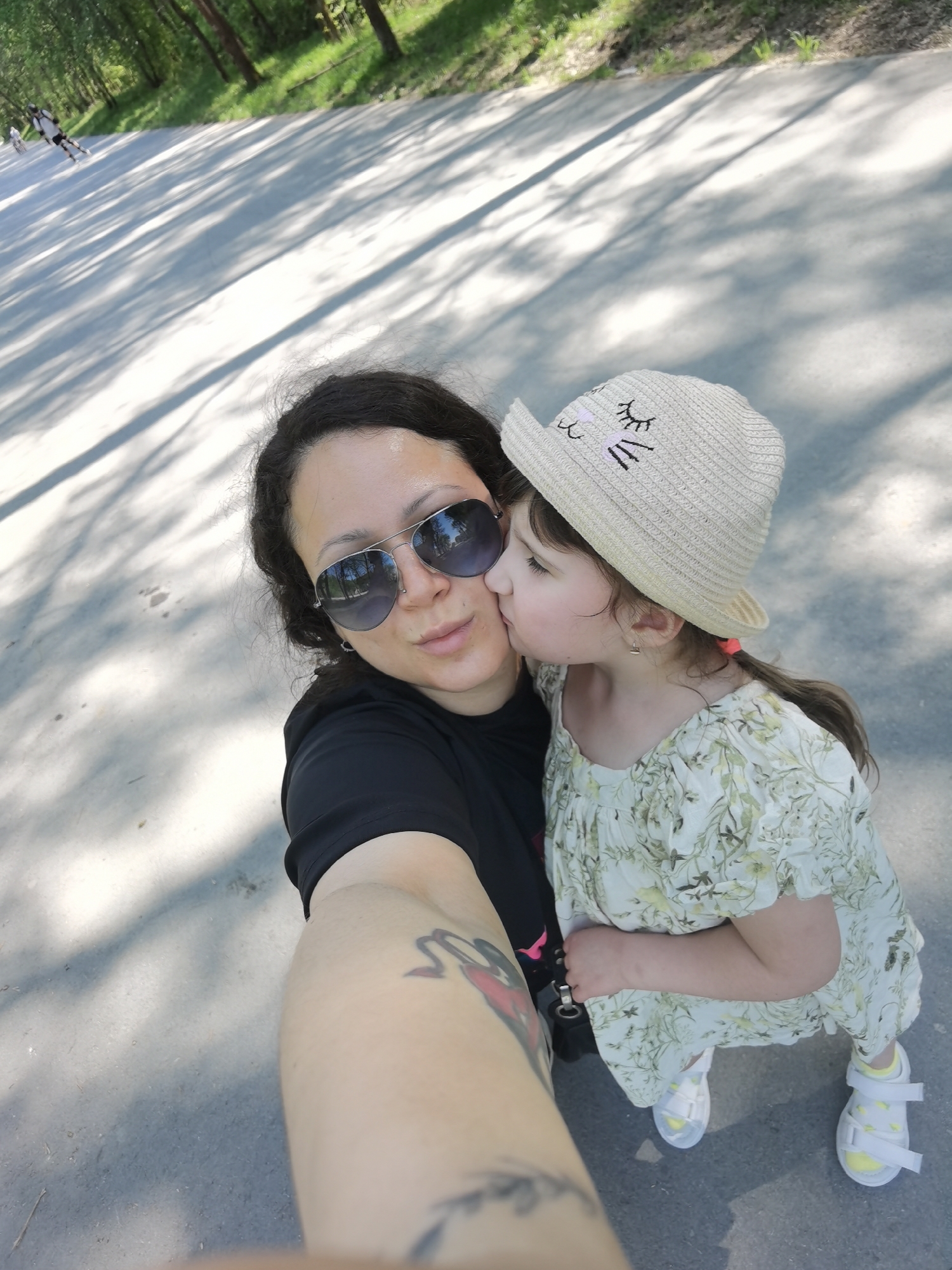 A selfie shows a woman and a child on a paved path in a park-like setting. The woman, wearing aviator sunglasses and a black t-shirt, holds the camera. Her left arm, visible, has a tattoo. A young girl wearing a floral dress and a cat-eared sun hat is kissing the woman on the cheek. The setting is outdoor, with trees and shadows visible on the path. People roller skating are in the background. The location is likely a park in Perm, Russia.