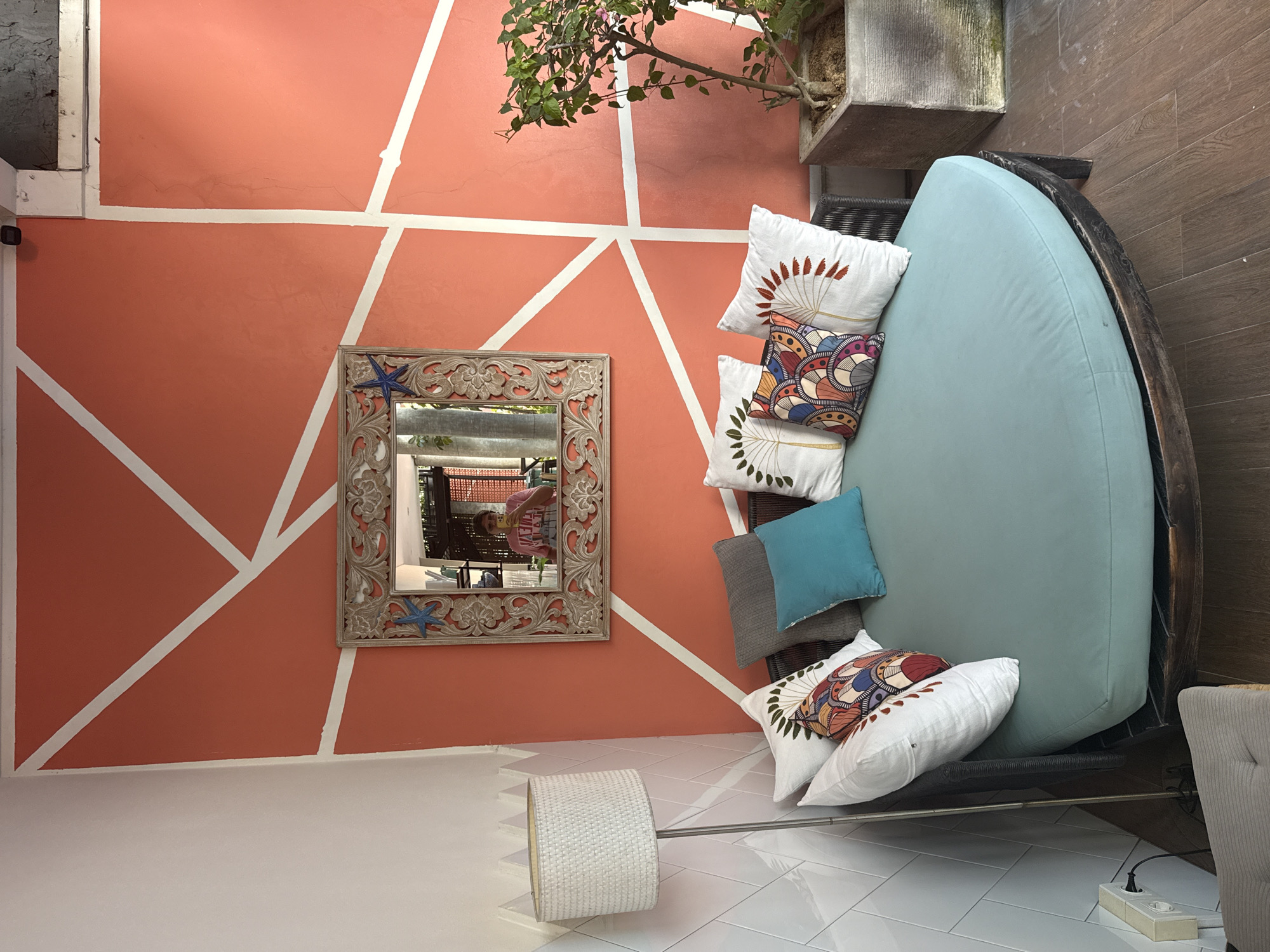 The image shows an interior space, likely a part of a residence or a hotel. The city is North Kuta, Indonesia. The walls are painted a salmon color with geometric white lines. A decorative mirror with a starfish motif hangs on the wall, reflecting a scene outside. A round daybed with a light blue cushion is arranged with several decorative pillows, including white pillows with various patterns and a plain teal pillow, along with a gray pillow. A potted plant sits adjacent to the daybed. In the foreground, there is a floor lamp with a white woven shade. The floor is covered in glossy white tiles. Part of a grey upholstered armchair is also visible on the right.