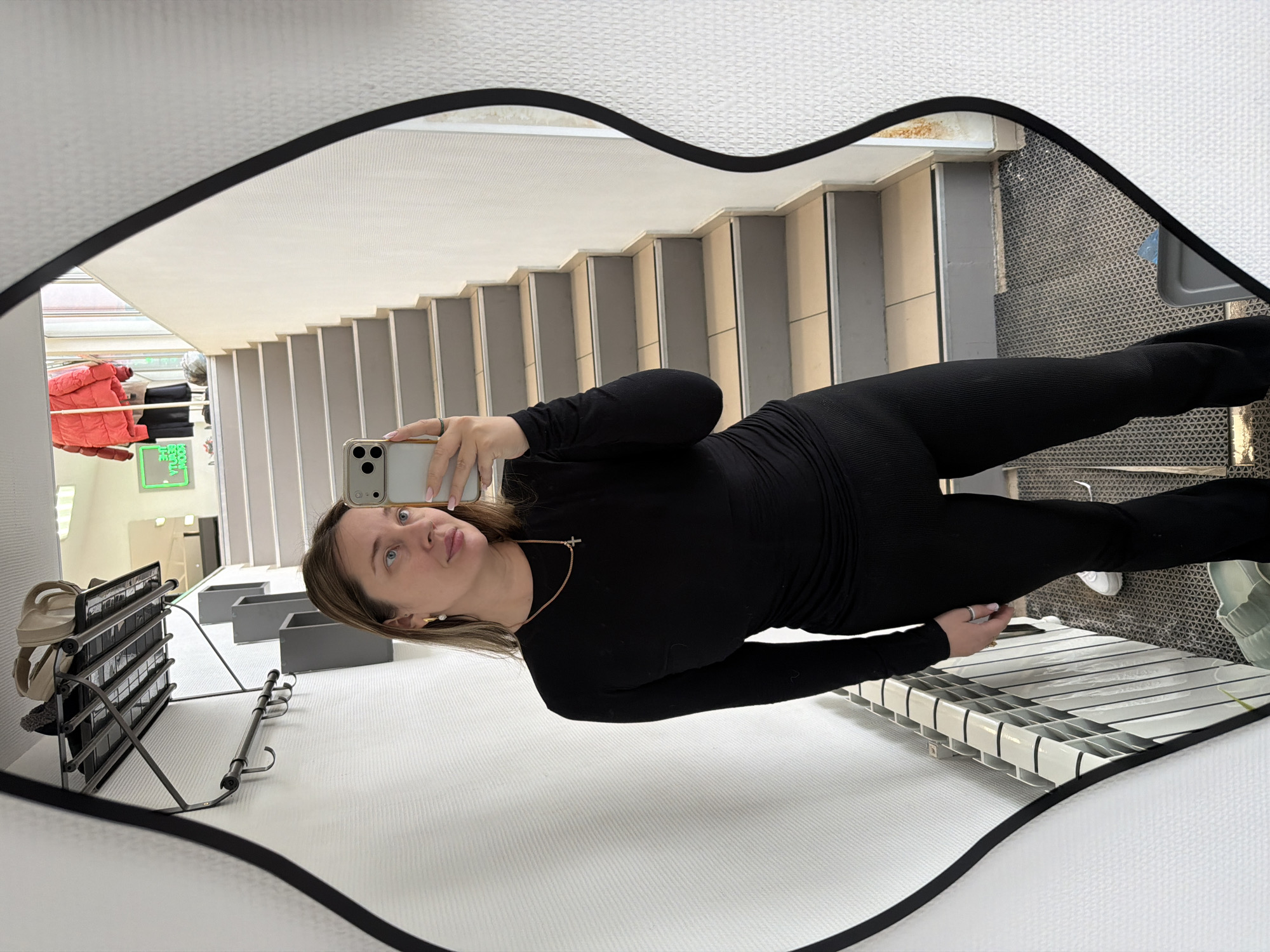 This image depicts a female subject taking a mirror selfie within an interior space, identified as being in Chaikovsky, Russia. The subject, a light-skinned individual with medium-length light brown hair, is wearing a form-fitting black long-sleeved top and black ribbed flared trousers. She is adorned with a gold chain featuring a cross pendant, gold hoop earrings with white pearls, and a ring on her right ring finger. Her left hand holds a light-colored smartphone, positioned to capture her reflection.
The environment reflected in the mirror features textured white wallpaper walls. To the immediate left, a black metal wall-mounted coat rack with a shelf holds a beige handbag and a dark, possibly fur-lined object. Below this, a white radiator is visible. The floor beneath the subject is covered by a gray patterned mat. Beyond, a flight of stairs with gray treads and light-colored risers ascends into the upper background. Further up the stairs, a bright red puffer jacket hangs on a visible clothing rack, and a partially obscured, reversed neon green sign (possibly displaying text like "THE ROOM") suggests a commercial retail establishment.