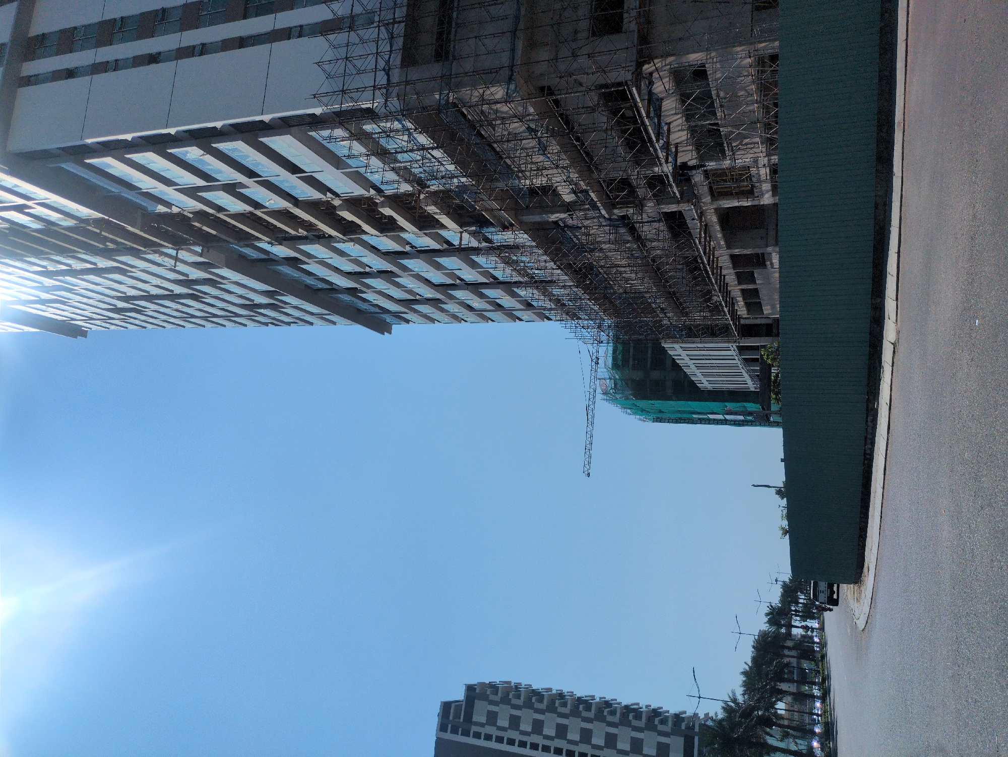 The image shows a street scene in South Quy Nhon Ward, Vietnam, featuring buildings under construction. A tall building with a grid-like facade occupies the left side of the frame, while another building under construction, enveloped in scaffolding, is positioned adjacent to a green fence on the right. A crane is visible in the middle ground, indicative of ongoing construction. The foreground consists of a paved street, and in the distance, there are trees and other buildings. The sky is clear and blue, suggesting a sunny day.