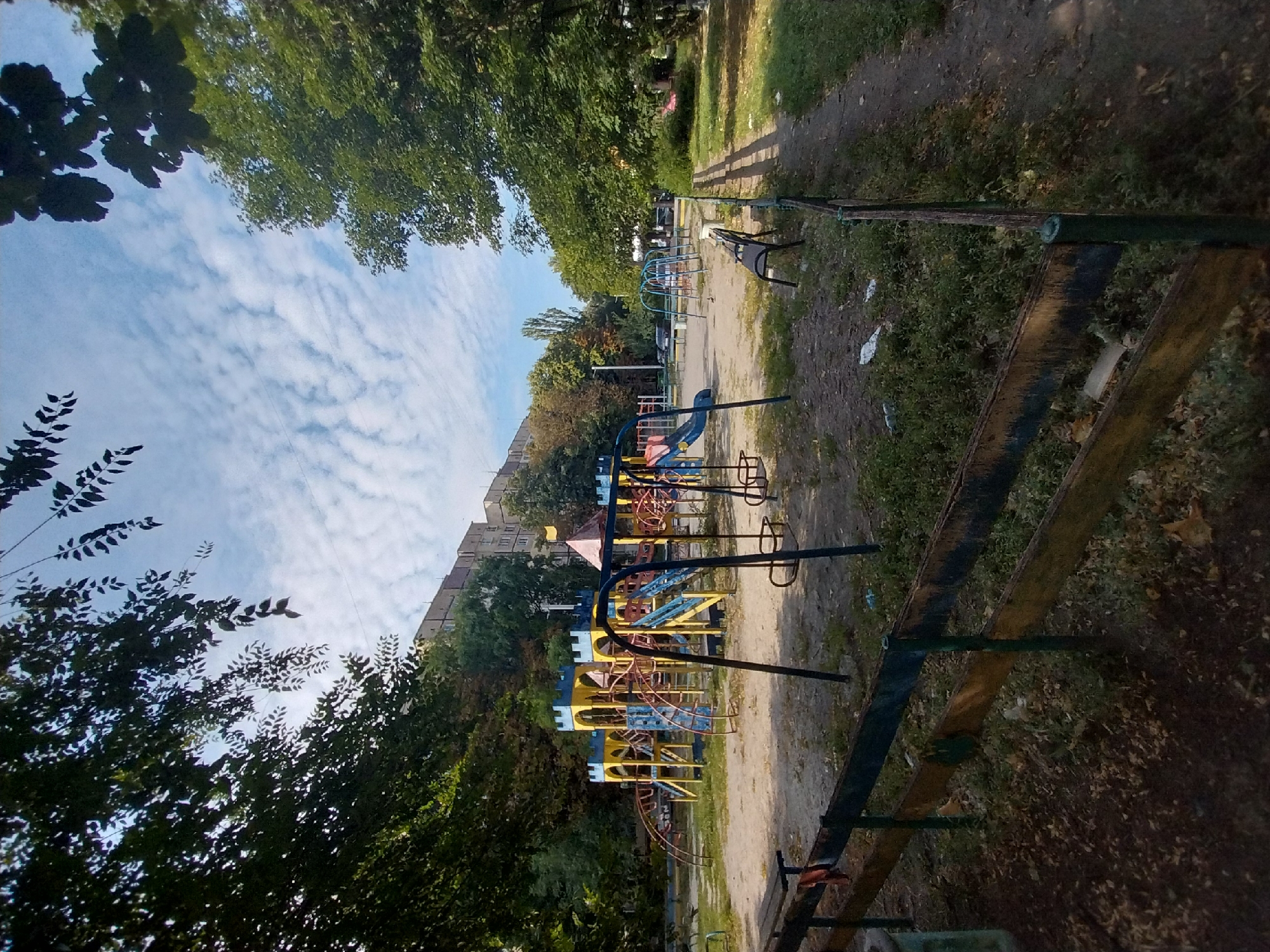 The image shows a playground in a residential area. The playground features a large, colorful structure resembling a castle with slides and climbing elements. There are also swings and benches. The playground is surrounded by trees and a low wooden fence. In the background, there are apartment buildings and parked cars. The sky is partly cloudy. The scene appears to be in Dnipro, Ukraine, based on common residential architecture and playground styles found in the region. No people are visible in the image. The environment is typical of an urban courtyard in Eastern Europe.