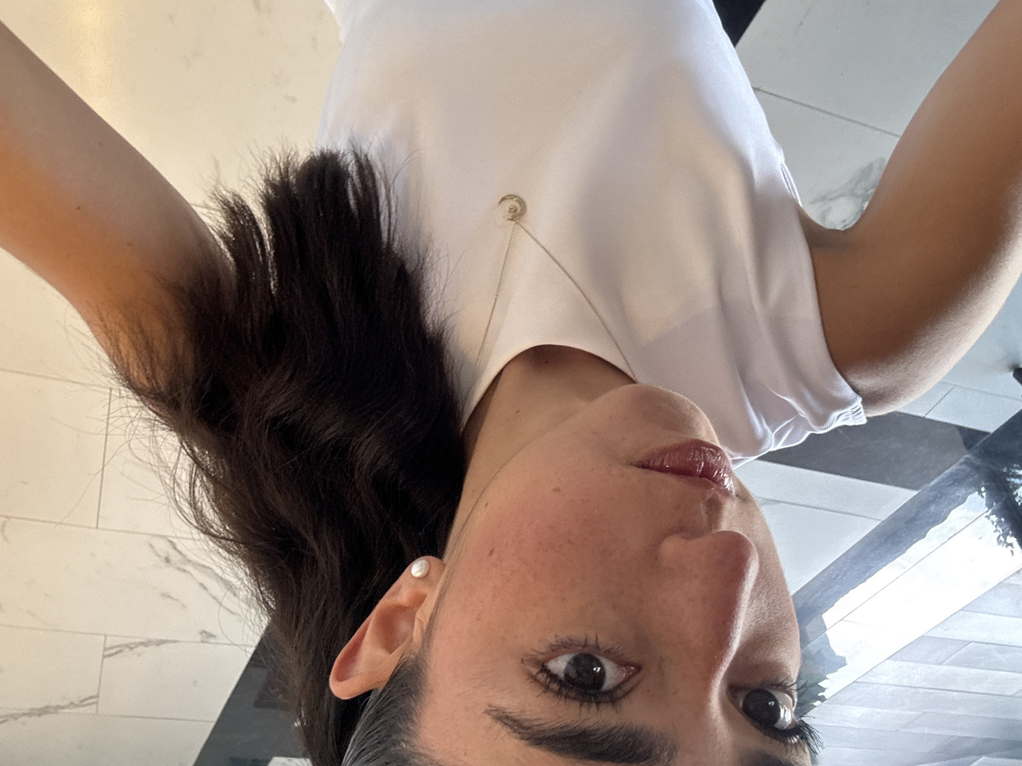 A woman with dark hair and a white t-shirt takes a selfie. She is wearing a pearl earring and a necklace with a pendant. The background is a black and white tiled floor. The location is Unknown, Unknown.
