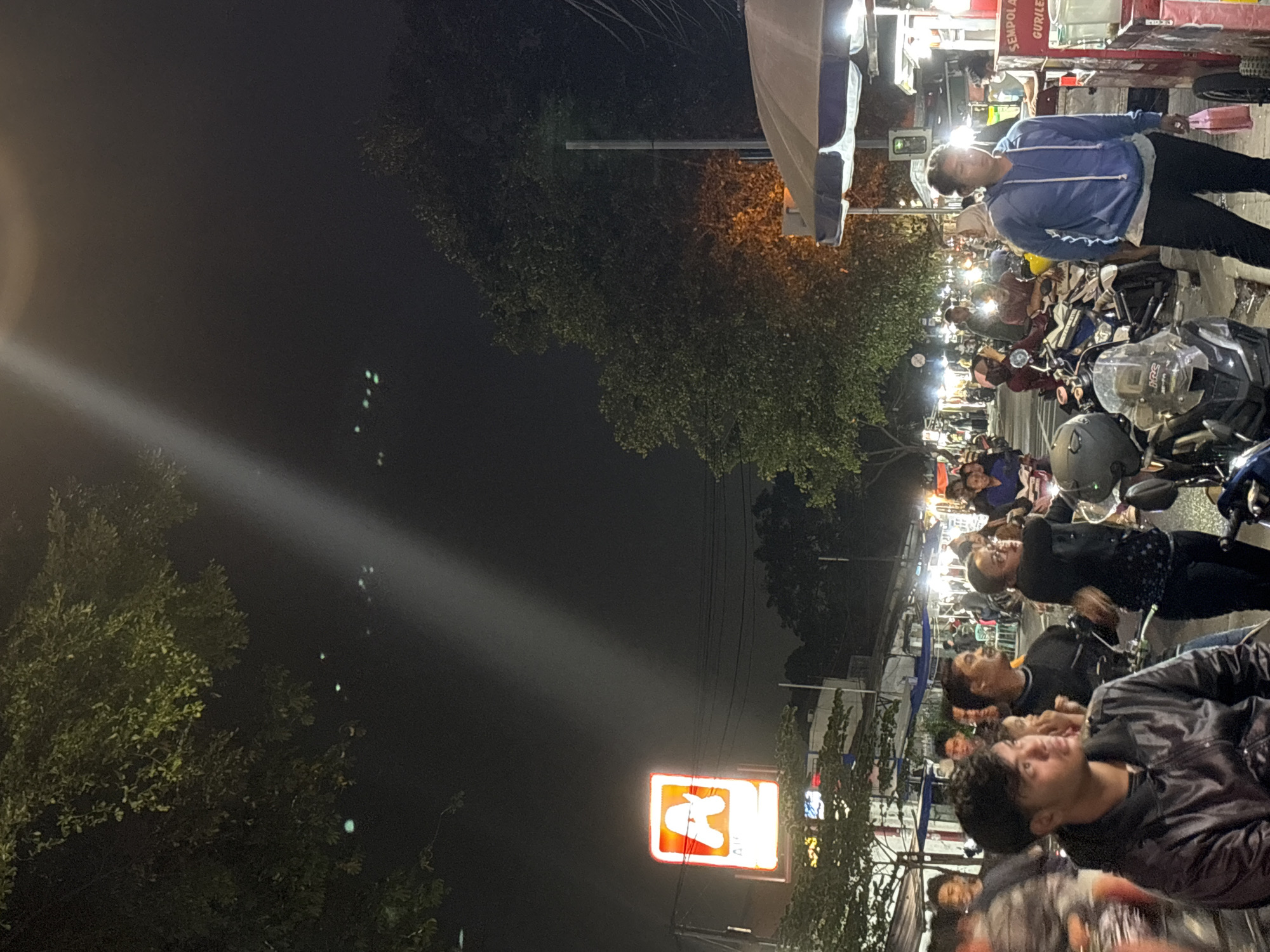 The image depicts a lively street scene at night in North Jakarta, Indonesia. Several people are gathered on the side of the road, some riding motorbikes, while others are standing and conversing. Many are illuminated by the bright street lights. Above the crowd, a brightly lit sign for "AlfMart" is seen. The sky is dark and the foliage around the street is partially visible, suggesting a partially residential area. A few food stalls are also present, with at least one person appearing to be purchasing something.