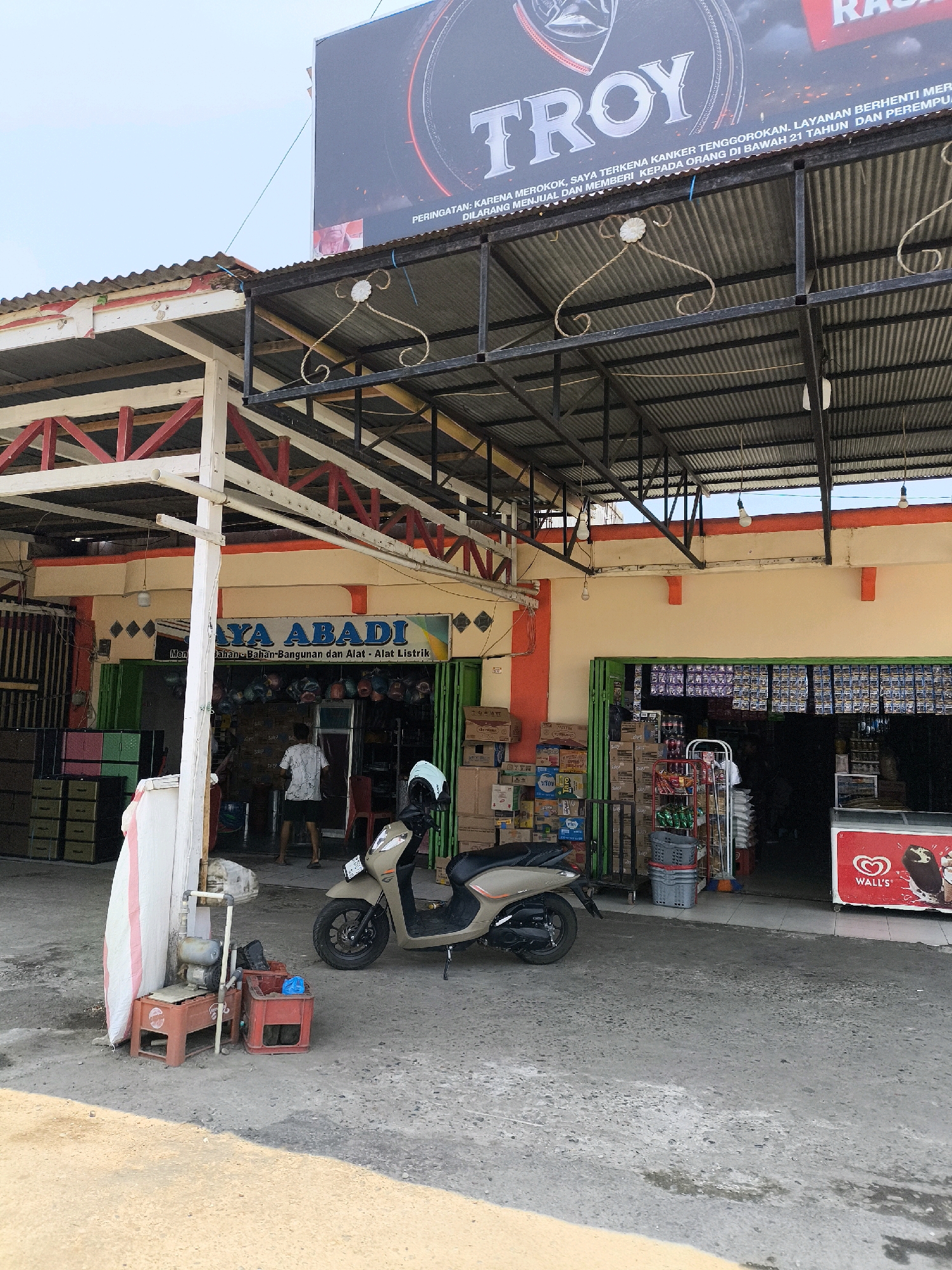 The image shows a street-level view of a storefront in Kwandang, Indonesia. The storefront is a shop named "JAYA ABADI" which sells building materials and electrical supplies. In front of the store, a beige scooter is parked, and a person is standing inside the shop. The shop is stocked with various goods, including boxes of products and a Wall's ice cream freezer. The storefront is covered by a corrugated metal roof supported by a metal structure. Above the storefront, a large billboard with the brand name "TROY" is visible. The ground in front of the shop is paved with concrete and dirt.