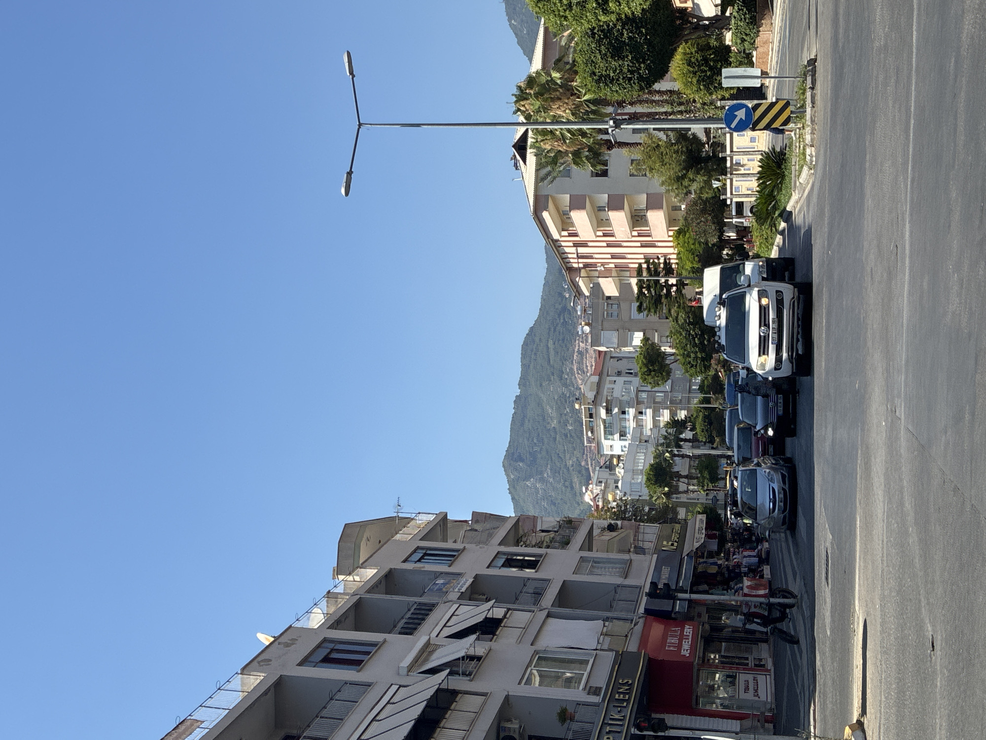 The image captures a street scene in Alanya, Turkey, on a clear, sunny day. In the foreground, a road with multiple lanes is visible, with several cars stopped at an intersection. A white Volkswagen SUV is prominent, with a license plate that appears to read "607 UJP 197". Behind it, other vehicles, including a silver sedan and a dark SUV, are also present.
On the left side of the street, a multi-story building with balconies and awnings lines the sidewalk. Several shops are situated on the ground floor, with signs indicating "OPTİK-LENS" and "FİBULA JEWELLERY." A delivery person on a scooter with a large red delivery box is seen near the shops.
On the right side of the intersection, another multi-story building with balconies, painted in pale yellow and cream, stands. Palm trees and dense green foliage are interspersed between the buildings, indicating a warm climate. In the background, rolling hills covered in vegetation rise, with some smaller buildings visible on their slopes. A tall street lamp with two lights is positioned near the right side of the road. A blue directional sign with a white arrow pointing to the right is also visible. The sky is a bright, cloudless blue.