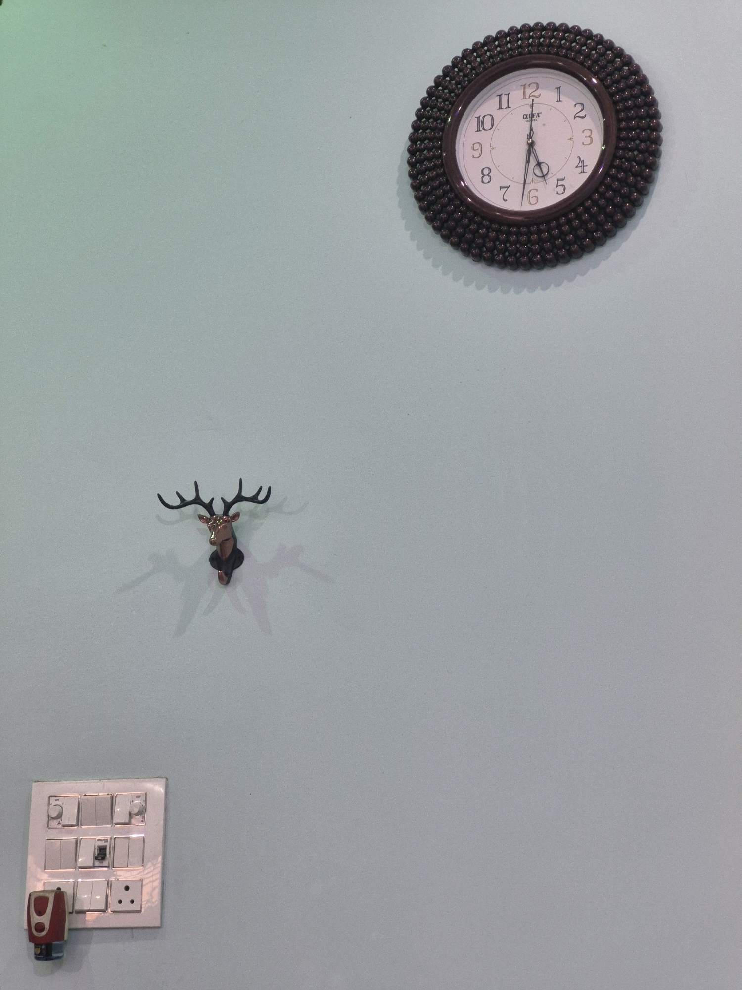 A wall-mounted clock with a dark, beaded frame is positioned on a light blue wall. The clock face has black Arabic numerals and hands. To the left of the clock, a decorative deer head with antlers is mounted on the wall. Below the deer head, a white electrical switchboard with multiple switches and a universal socket is installed. A small red and black plug-in device is attached to the bottom of the switchboard. The lighting suggests a room environment, and there are subtle shadows cast by the objects on the wall. There is no indication of the city of Laloni, India in the image.