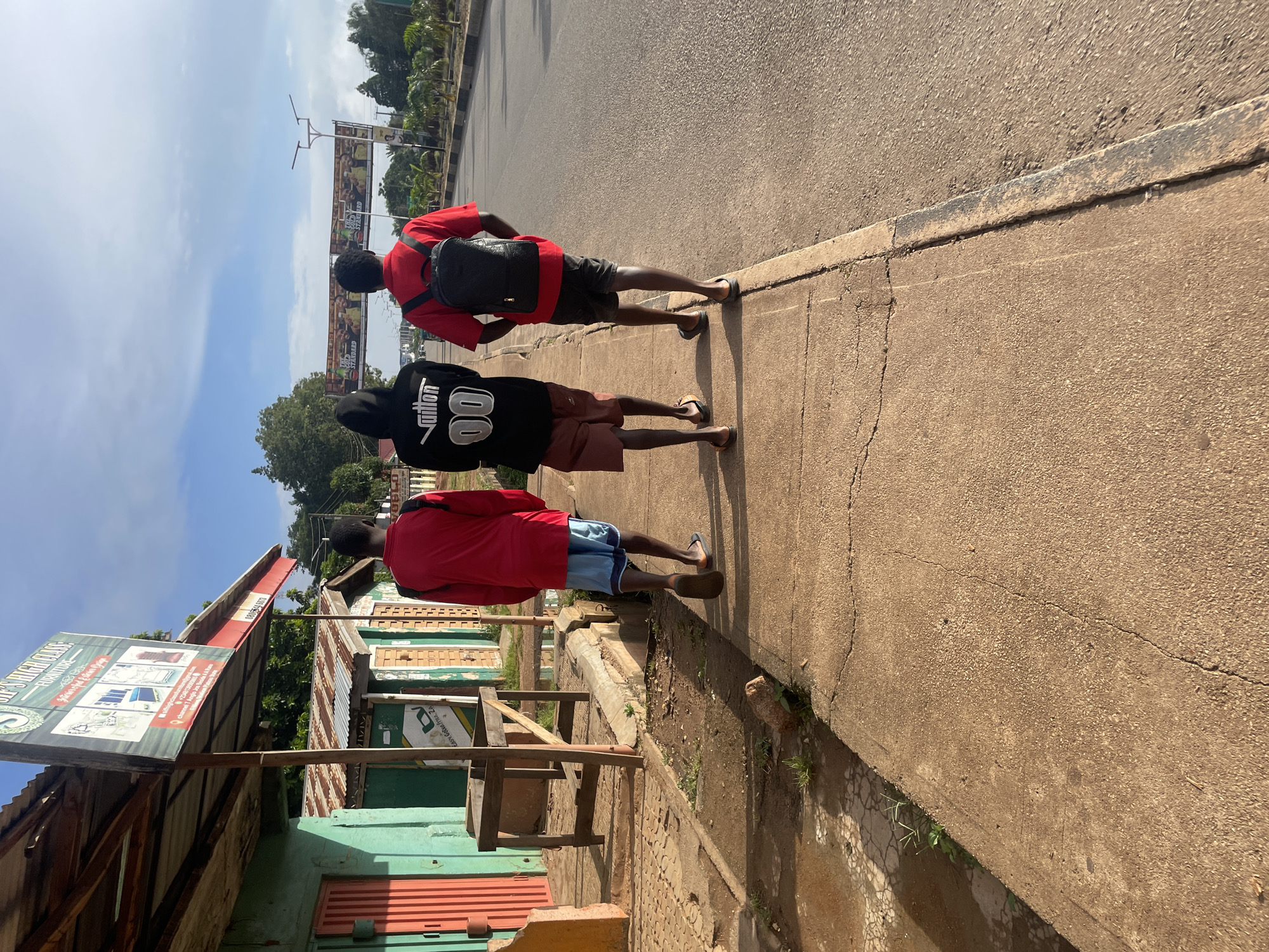 Three young men are walking away from the camera on a paved sidewalk next to a road. The man on the left is wearing a red long-sleeved shirt and blue patterned shorts. The man in the middle is wearing a black hooded sweatshirt with white lettering and designs, and brown shorts. The man on the right is wearing a red long-sleeved shirt and black shorts, and is carrying a black backpack.
They are walking past a row of small buildings with corrugated metal roofs. The building on the far left has a green facade and a red door. A sign for "SIP'S HIGH CLASS FURNITURE" is visible above the buildings. The road to the right appears to be a multi-lane highway with streetlights. In the distance, a banner with images of people and text is stretched across the road. The scene appears to be in Jos, Nigeria, as indicated by the presence of "SIP'S HIGH CLASS FURNITURE" which is a known business in Jos. The time of day appears to be late afternoon, with long shadows cast by the setting sun. The sky is partly cloudy.