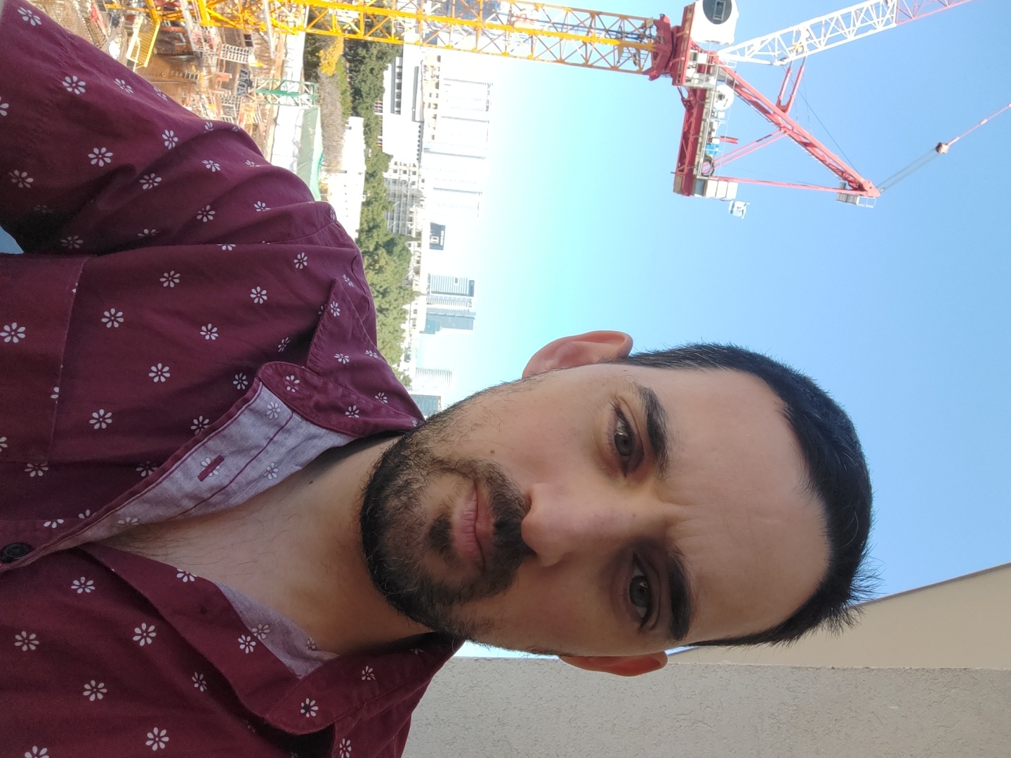 A close-up selfie captures a man against an urban backdrop dominated by a construction site and clear blue sky. The man, with dark hair, a beard, and dark eyes, wears a dark red collared shirt adorned with small white floral patterns. His expression is neutral to slightly serious as he looks directly at the camera. The image is tilted, presenting a dynamic, informal perspective.
Behind him, a bustling construction area is visible, featuring large yellow and red cranes reaching towards the sky. In the mid-ground, a mix of completed city buildings and structures still under development can be seen, interspersed with green trees. The prominent feature in the upper background is a large red and white crane, contrasting sharply with the bright, cloudless blue sky, indicating a sunny day. The scene suggests an active urban environment, consistent with the provided location context of Tel-Aviv, Israel, highlighting ongoing development. There is no visible text in the image.