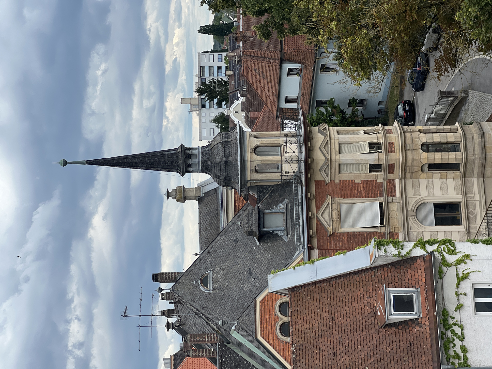 This is an eye-level, high-angle shot of a cityscape in Straubing, Germany, under a cloudy sky. The focal point is a narrow, dark spire topping a detailed, multi-story building with ornate architecture. The spire is a dark, segmented cone with a small finial at its peak. Below the spire, the building features bay windows with arched panes and decorative molding. Red tiled roofs dominate the foreground and middle ground, with several chimneys visible. A light blue building with a red tiled roof sits to the right of the spire building. A dark-colored sedan and a blue SUV are parked on a paved street that curves through the scene, bordered by a metal fence and trees. In the distance, a multi-story apartment building with solar panels on its roof is visible, along with a line of trees. A solitary bird is flying high in the overcast sky.