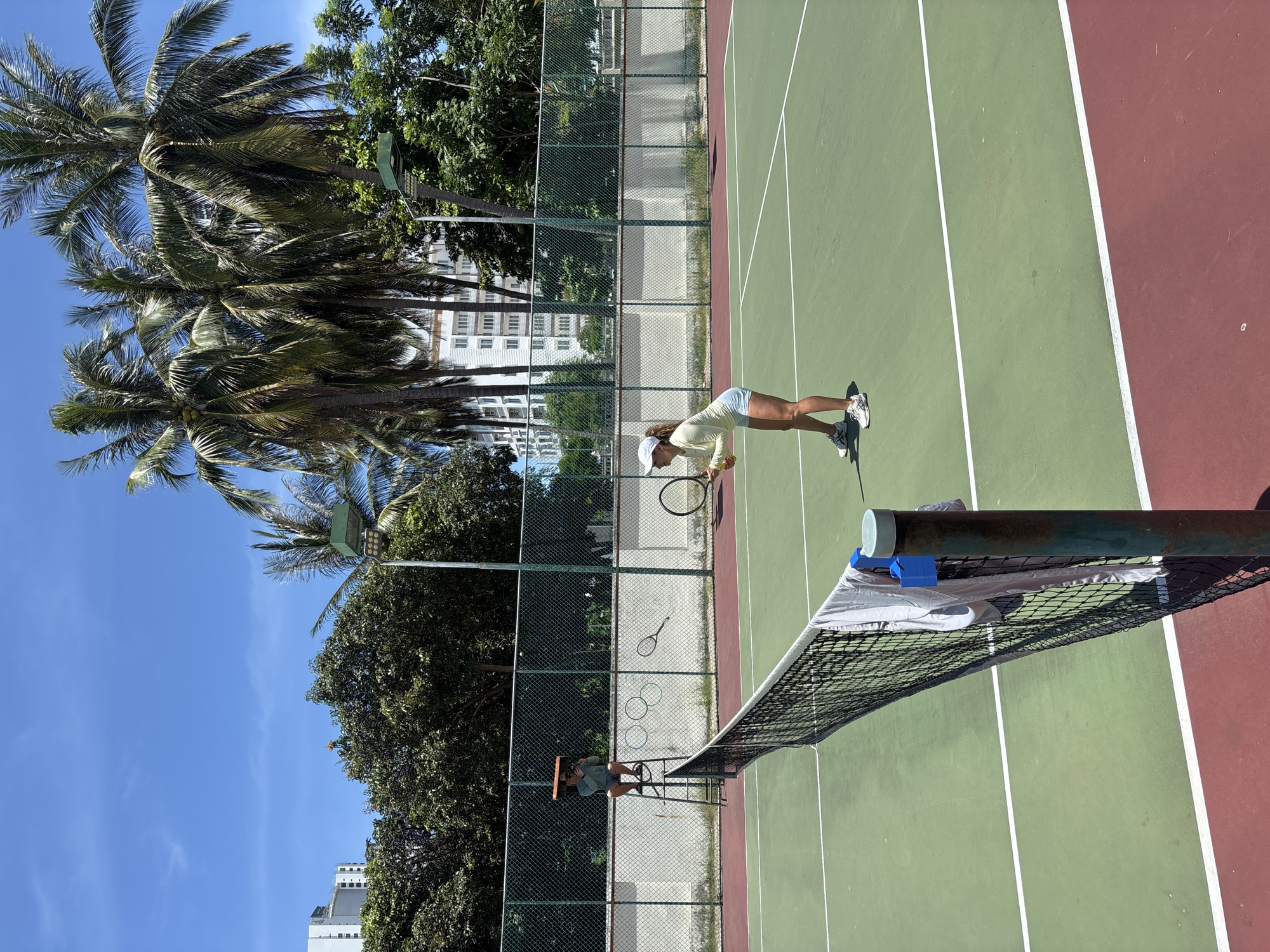 This image shows a tennis court located in Nha Trang, Vietnam. The court has a green surface with red outer boundaries and a net dividing it in half. In the foreground, a towel is draped over the net. On the far side of the net, a woman is standing on the court, holding a tennis ball and a racket, appearing to be in the middle of a game. Behind her, palm trees and a tall building are visible, suggesting an urban coastal setting. On the near side of the net, a person sits on a raised umpire's chair, observing the game. The sky is clear and blue, indicating a sunny day.