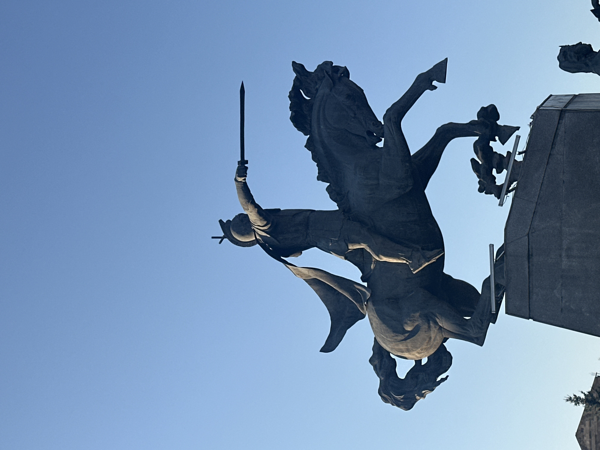 A low-angle, full shot captures a large bronze equestrian statue against a clear blue sky. The statue depicts a warrior wearing a helmet and a flowing cloak, mounted on a rearing horse. The warrior is holding a sword outstretched in their right hand, pointing horizontally to the right. Below the horse, a smaller sculptural element representing figures or mythological creatures is visible. The statue stands on a substantial stone pedestal. In the lower left corner, a portion of a building with a pointed roof and a dark evergreen tree are visible. The scene appears to be outdoors, with the sunlight casting shadows on the statue, indicating it is likely daytime. The location is identified as Gyumri, Armenia.