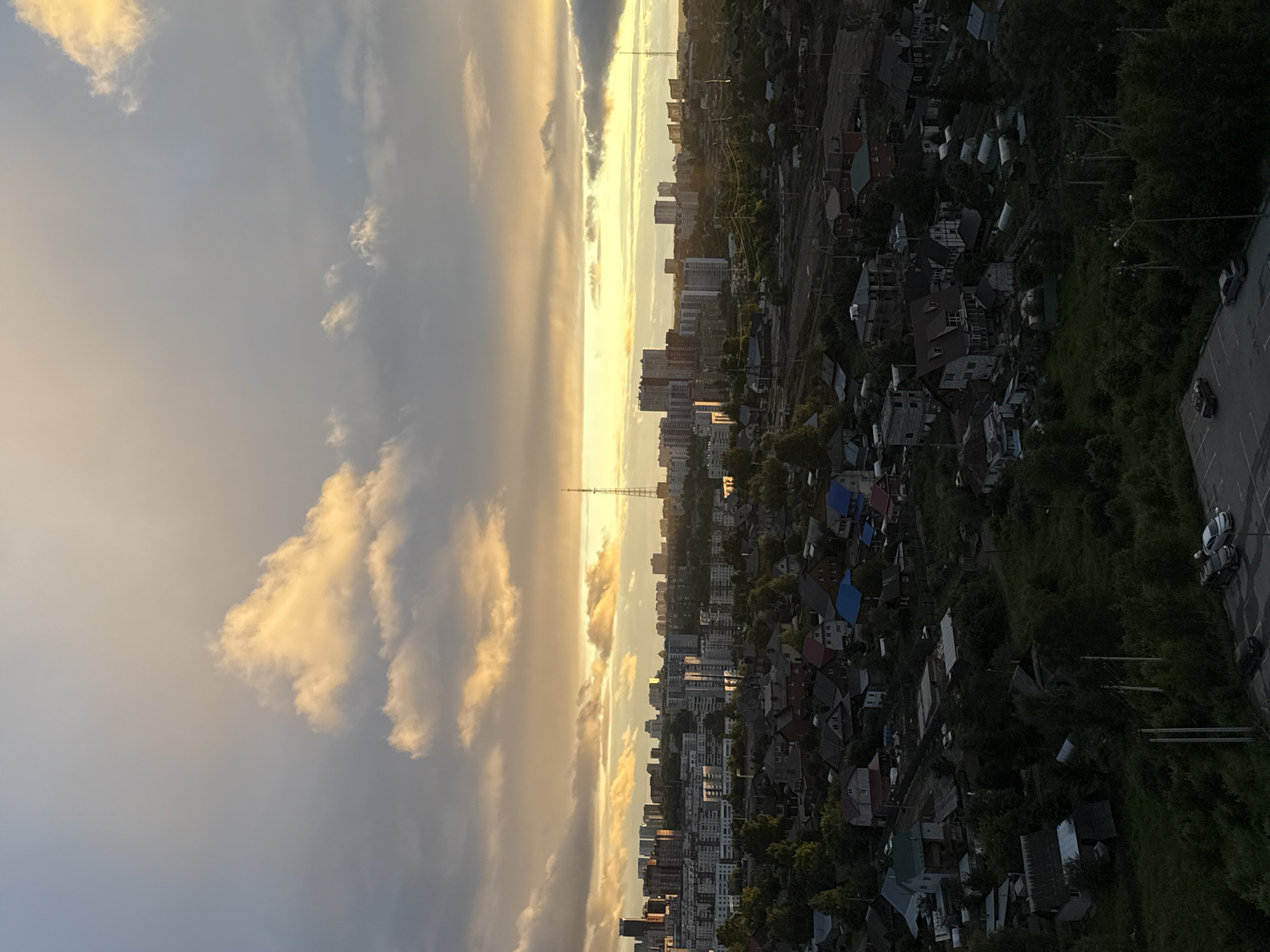 This is an aerial, high-angle, full shot of a city skyline in Perm, Russia, at sunset. The sky is filled with dramatic, illuminated clouds in shades of yellow and orange, contrasted with darker, grey clouds. The sun is setting on the horizon, casting a warm glow over the cityscape.
In the background, a dense urban area with numerous apartment buildings of varying heights and architectural styles is visible. Some buildings are modern, with glass facades, while others appear older and more traditional. A prominent radio tower stands tall among the buildings on the right side of the frame.
The middle ground features a transition from the urban core to a more suburban or semi-urban area. This section includes smaller residential buildings, possibly houses and townhouses, interspersed with trees and vegetation. Some buildings have distinct colored roofs, such as blue and red.
The foreground consists of a densely wooded area with numerous private houses, gardens, and greenhouses. The houses are of various sizes and designs, with some appearing to be of brick construction and others with darker siding. Trees and foliage are abundant, creating a sense of a greener, less developed area. A few cars are visible parked on a paved surface in the lower right corner of the image. The overall scene depicts a city at dusk, with the warm light of the setting sun illuminating the sky and casting long shadows over the landscape.