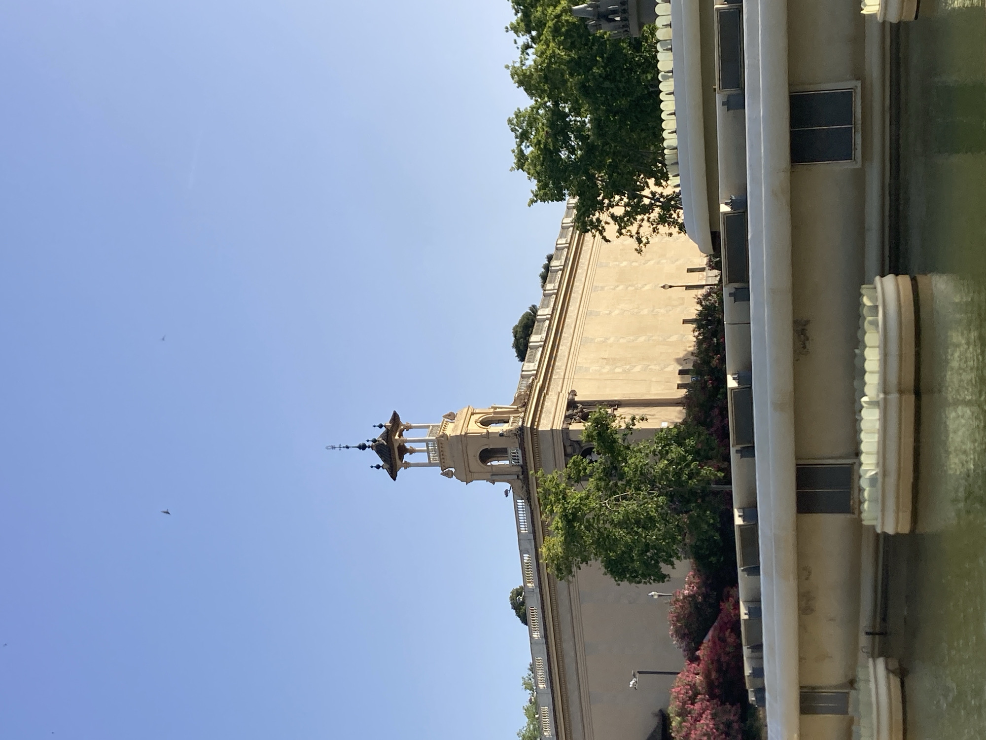 The image depicts a clear blue sky above a large, pale yellow building with a prominent bell tower featuring a decorative roof. The building has a long, ornate balustrade running along its upper edge. In the foreground, there is a body of water with a series of decorative, tiered fountains. Lush green trees and vibrant pink bushes are visible around the building and in front of it. The scene appears to be an exterior shot taken in daylight, likely in a public space. The architecture and fountains suggest a location within Barcelona, Spain, possibly the Palau Nacional on Montjuïc hill, which houses the Museu Nacional d'Art de Catalunya and features similar water features and a distinctive tower. There are no people visible in the frame.
