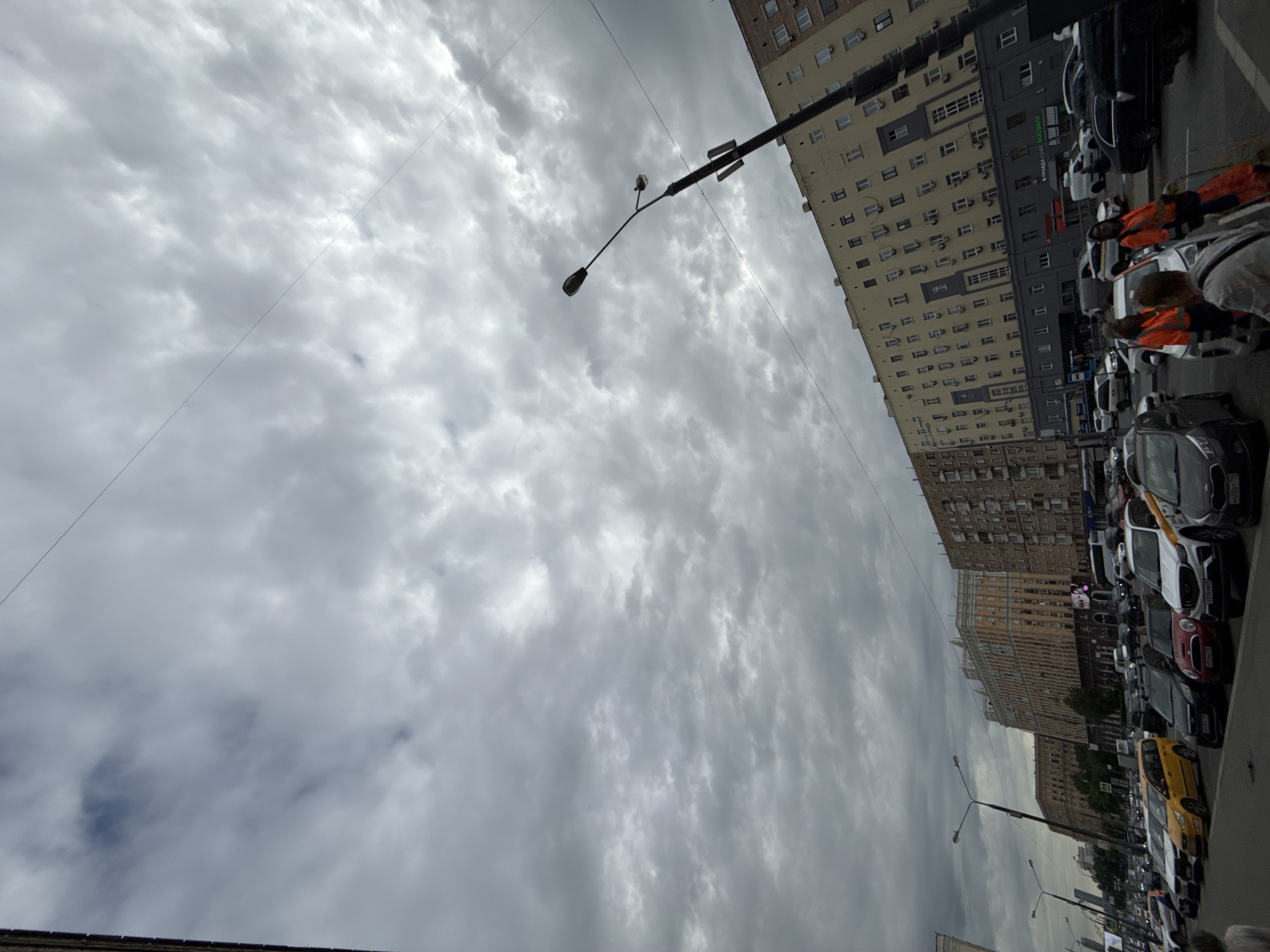 The image shows a street scene in Moscow, Russia. The view is directed upwards, capturing a cloudy sky occupying the majority of the frame. Several tall buildings line the right side of the image, displaying a variety of architectural styles and colors, ranging from beige to grey. A row of parked cars is visible along the street, with a few people wearing orange safety vests present. Streetlights are also interspersed along the road.