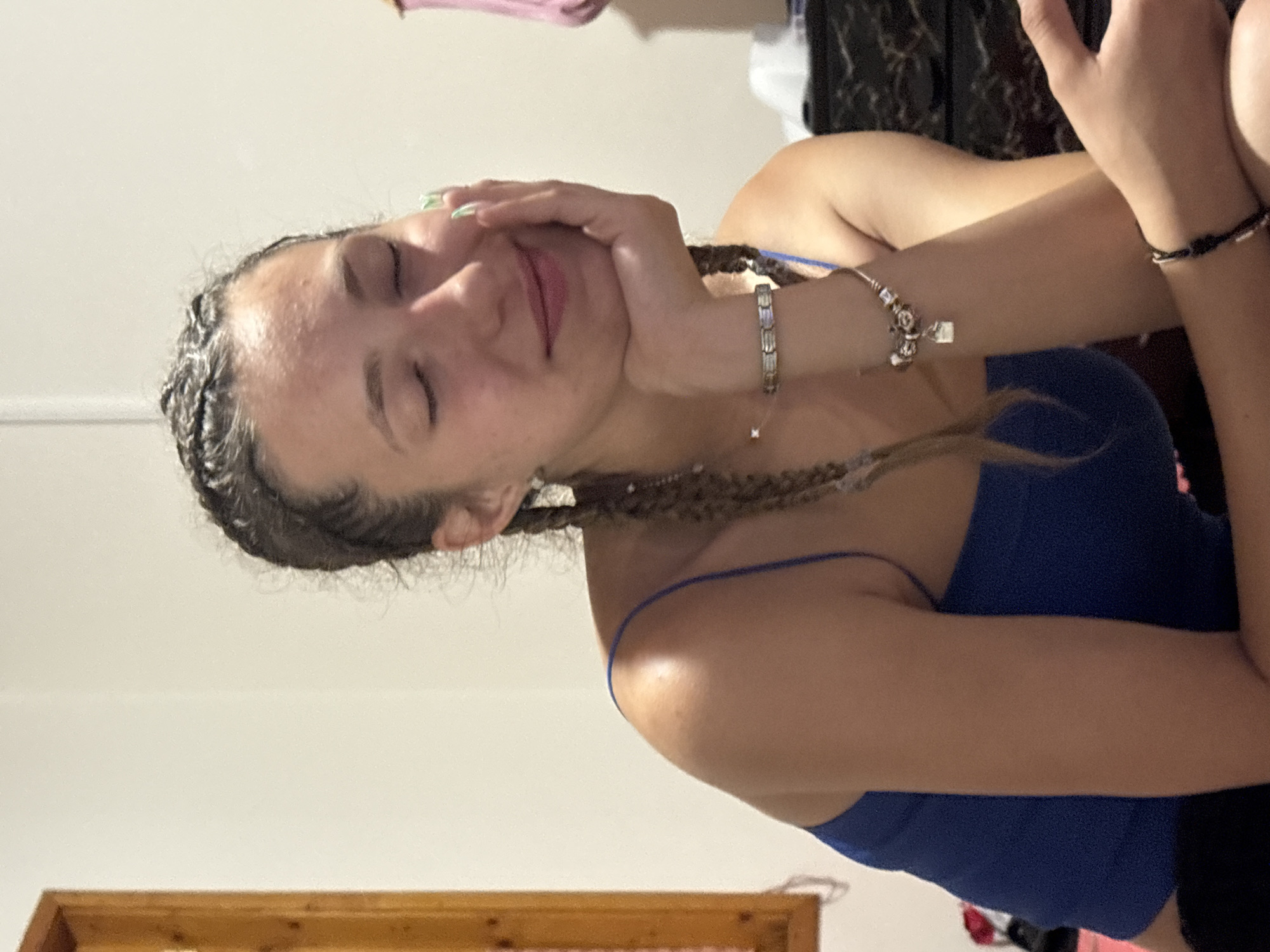 A young woman with her eyes closed is resting her head on her hand, supporting her chin. She has her hair in braids and is wearing a blue tank top. She has bracelets on her wrist and green painted fingernails. The background is a plain wall, with a wooden framed object on the left side of the image. This appears to be a portrait taken indoors, likely in a residential setting. No specific details suggest a location within Varna, Bulgaria.