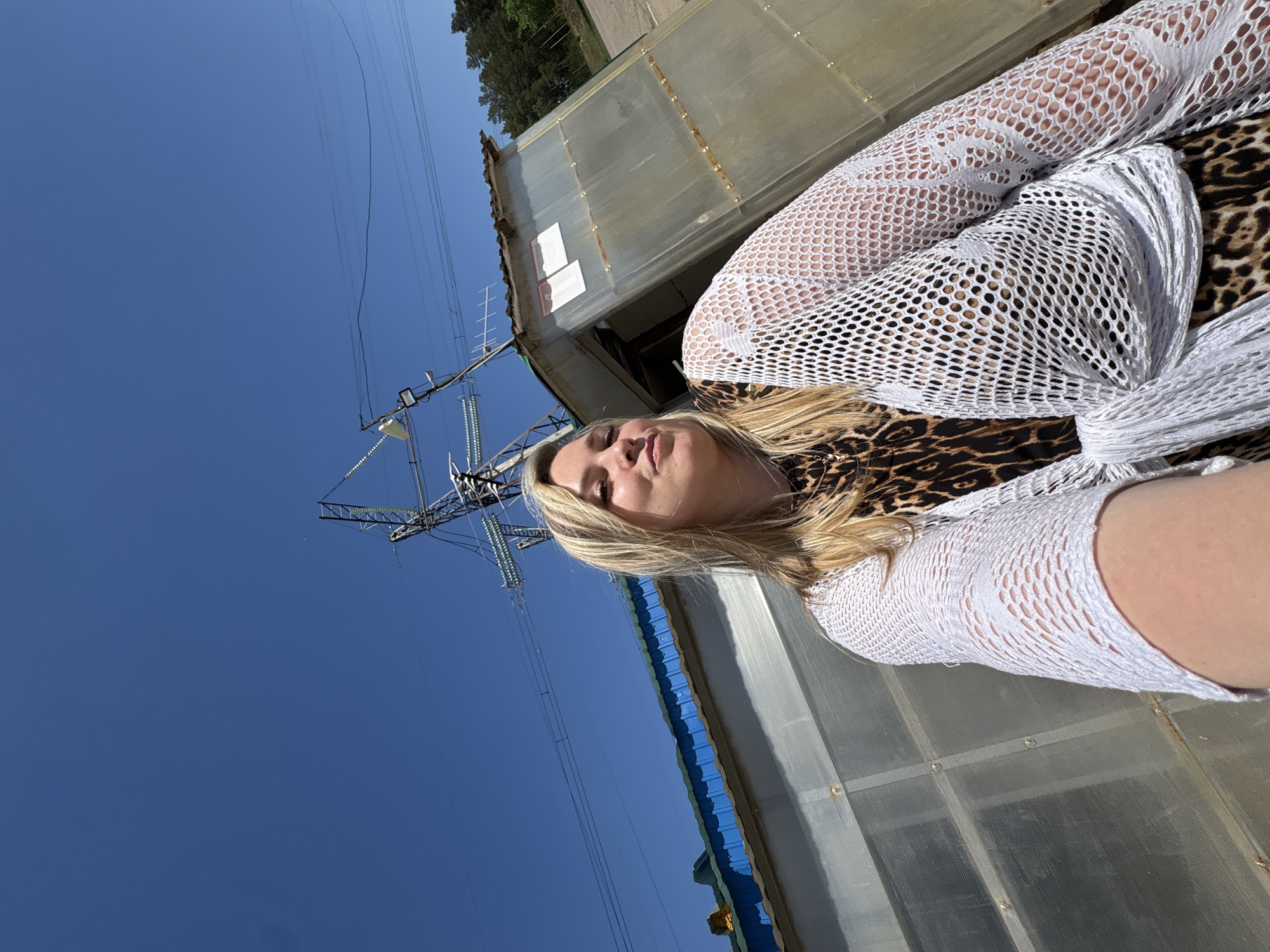 A woman with blonde hair is lying down, wearing a white, fishnet-like garment over a leopard-print top. A large, industrial-looking structure with a grey and blue exterior is behind her. In the background to the left, a power line tower stands against a clear blue sky. The location appears to be in an outdoor setting, possibly near an industrial area. The city of Noviy, Russia is not discernible from the image.
