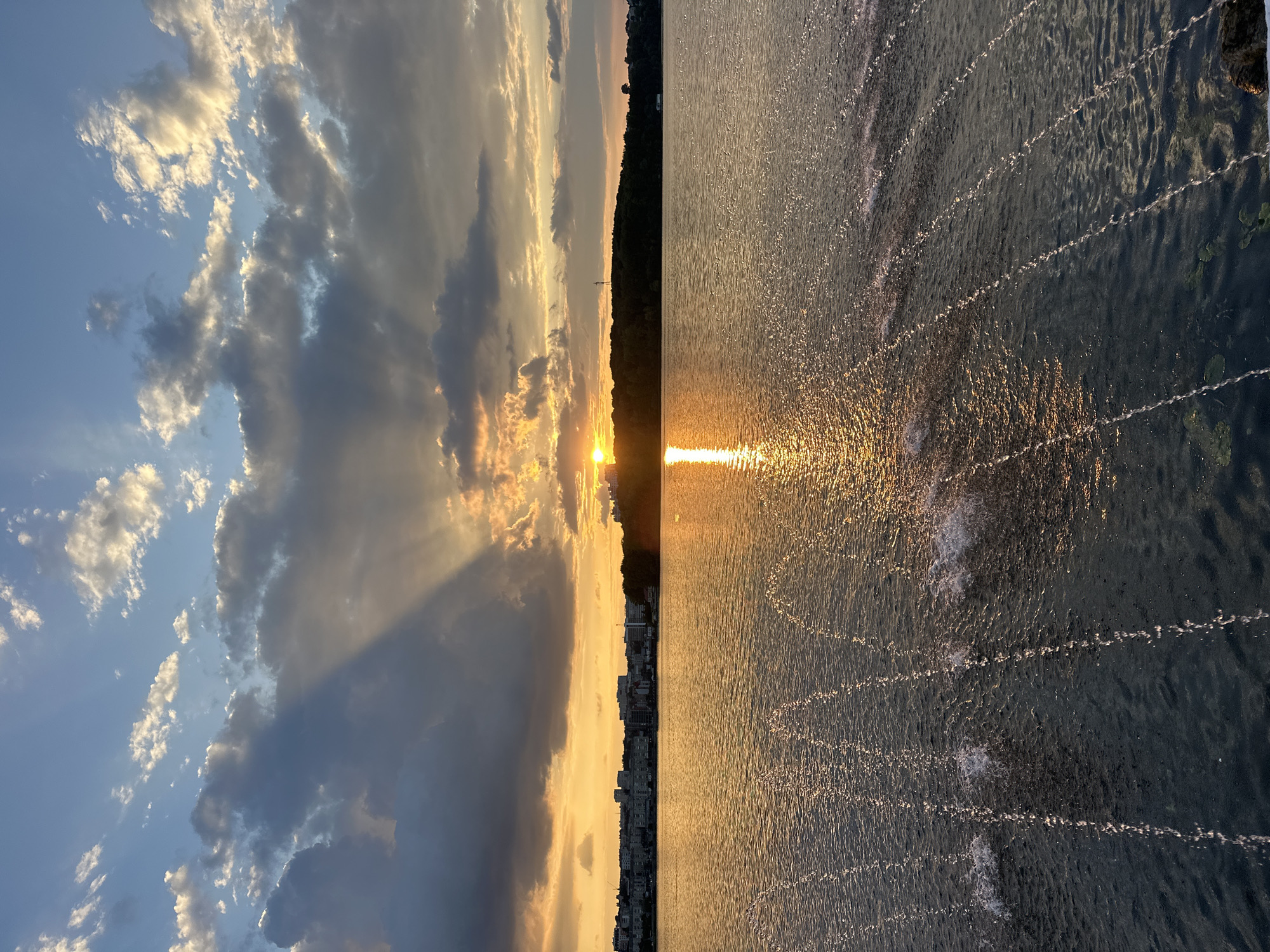 This is a photograph taken at sunset over the Ternopil Pond in Ternopil, Ukraine. The sun is low on the horizon, casting a golden glow across the water and sky. Dramatic rays of sunlight pierce through dark, heavy clouds, creating a striking contrast. In the distance, a cityscape with buildings lines the far bank of the pond, backed by a dense treeline. The foreground features a water fountain, with streams of water arcing and splashing into the pond, their surfaces catching the warm light of the sunset. The water is calm, with gentle ripples and the reflections of the sky and surrounding landscape visible. The overall scene is one of natural beauty and urban integration at the close of day.
