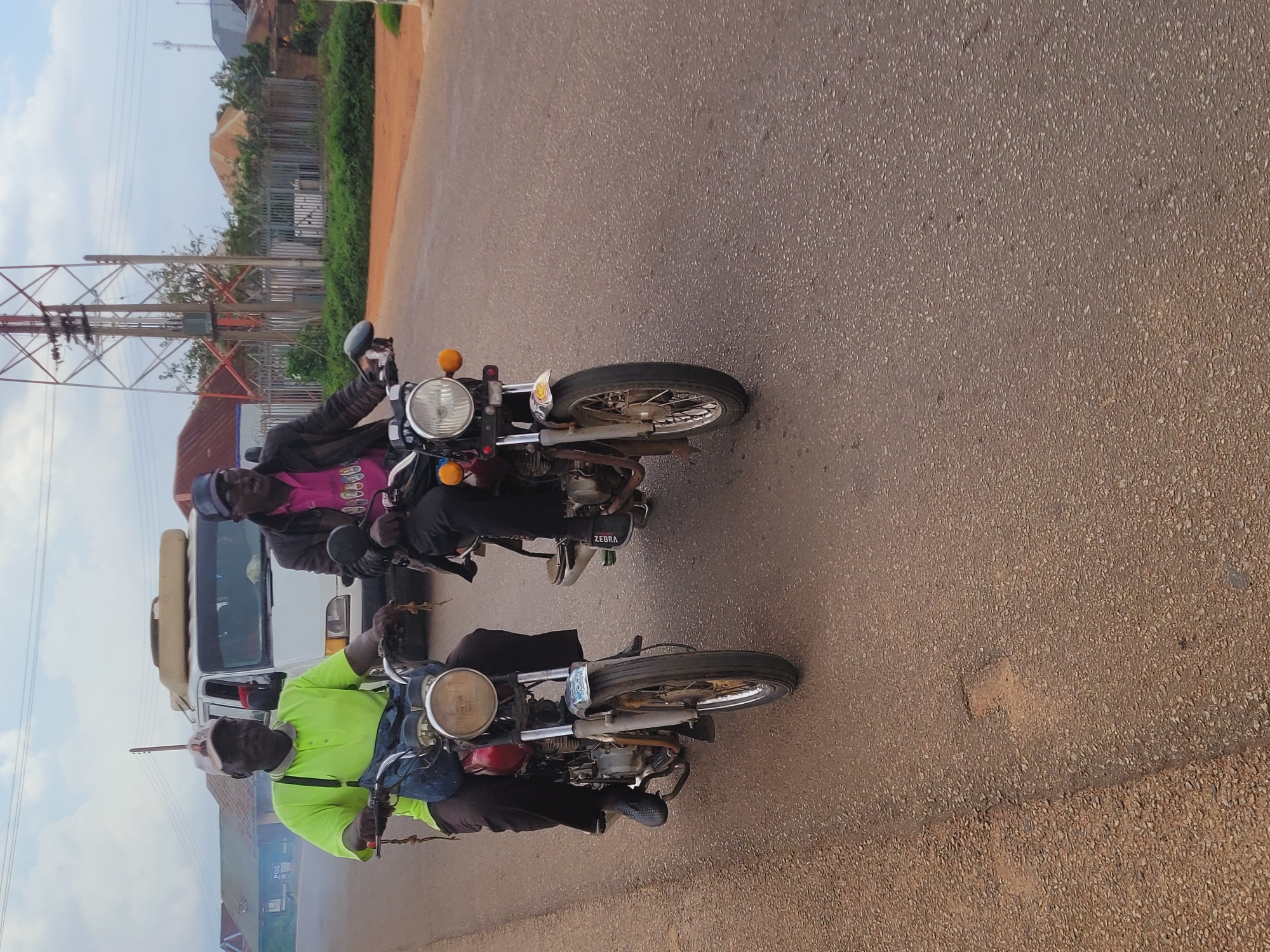 Two men ride motorcycles on a paved road in Du, Nigeria. The man in the foreground, on the left, wears a bright green polo shirt, dark pants, and a baseball cap. He is on a red motorcycle and has a blue bag slung over his shoulder. The man behind him, on the right, wears a dark jacket over a pink shirt and a cap. He is on a darker colored motorcycle. A white van is visible behind the man in green. The road is asphalt and appears to have some potholes. In the background, there are buildings with corrugated metal roofs and a large telecommunications tower. The sky is cloudy.