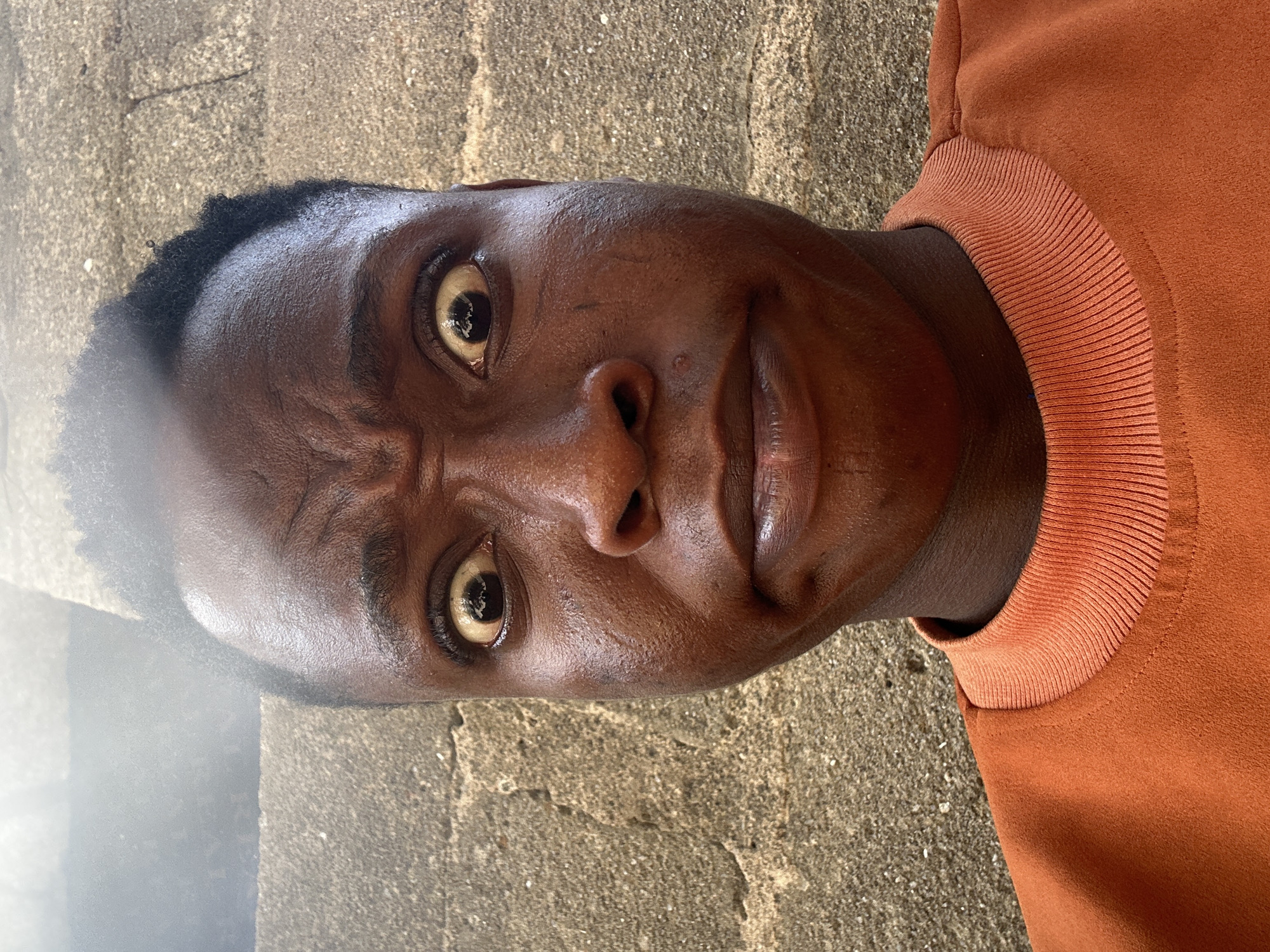 A medium shot of a dark-skinned man wearing an orange sweatshirt, taken outside on a stone surface. The man is close to the camera, looking directly at it with furrowed brows and a slight smirk. The location is in the city of Unknown, Unknown.