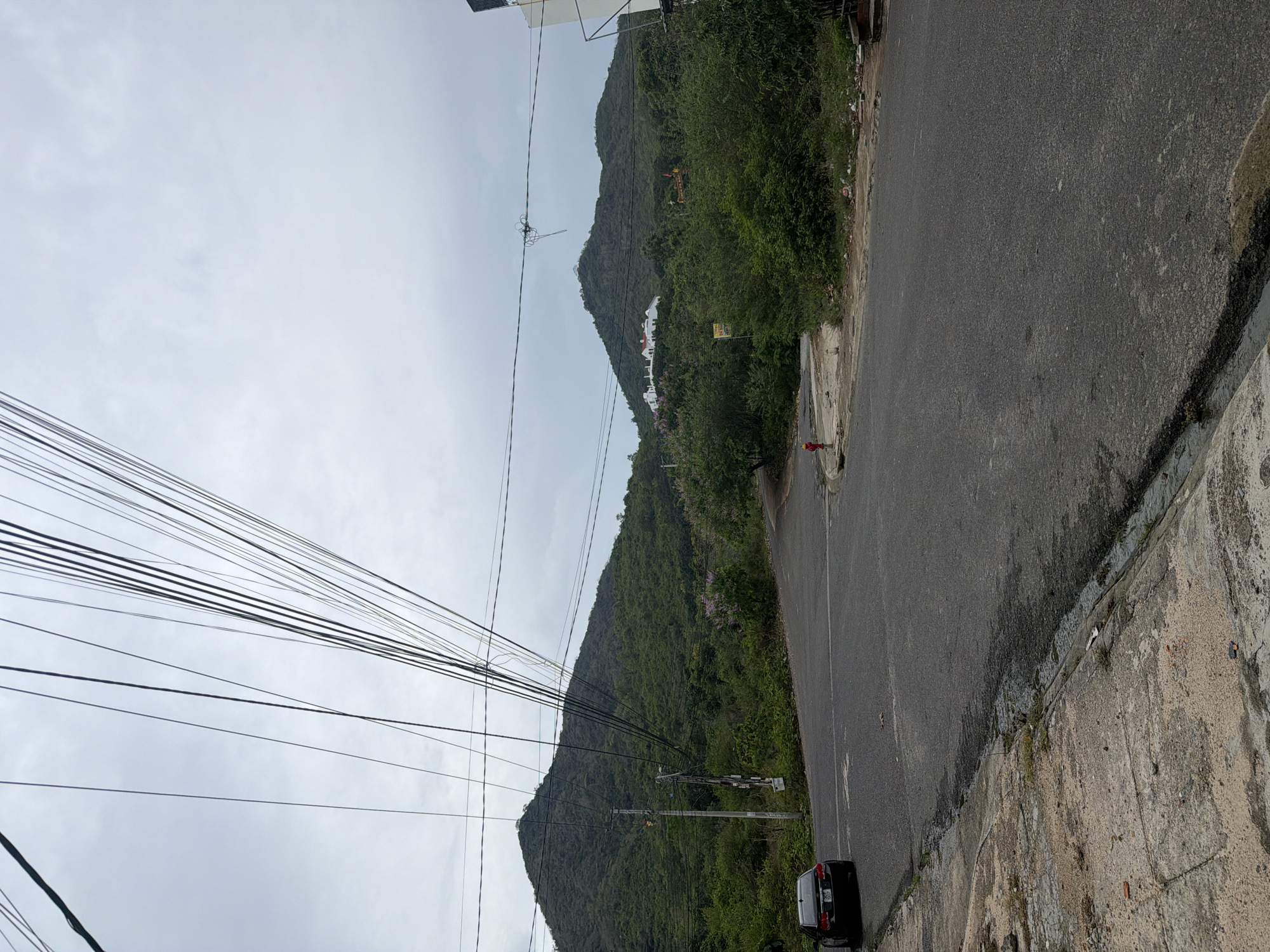The image depicts a street scene in Bac Nha Trang Ward, Vietnam. A paved road runs alongside a concrete curb and a wall. Lush green hills covered in dense vegetation rise in the background. Power lines crisscross the sky. A black car is parked near the edge of the road. A red fire hydrant is visible. A utility pole stands on the side of the road. Buildings are visible on the hills. The sky is overcast.