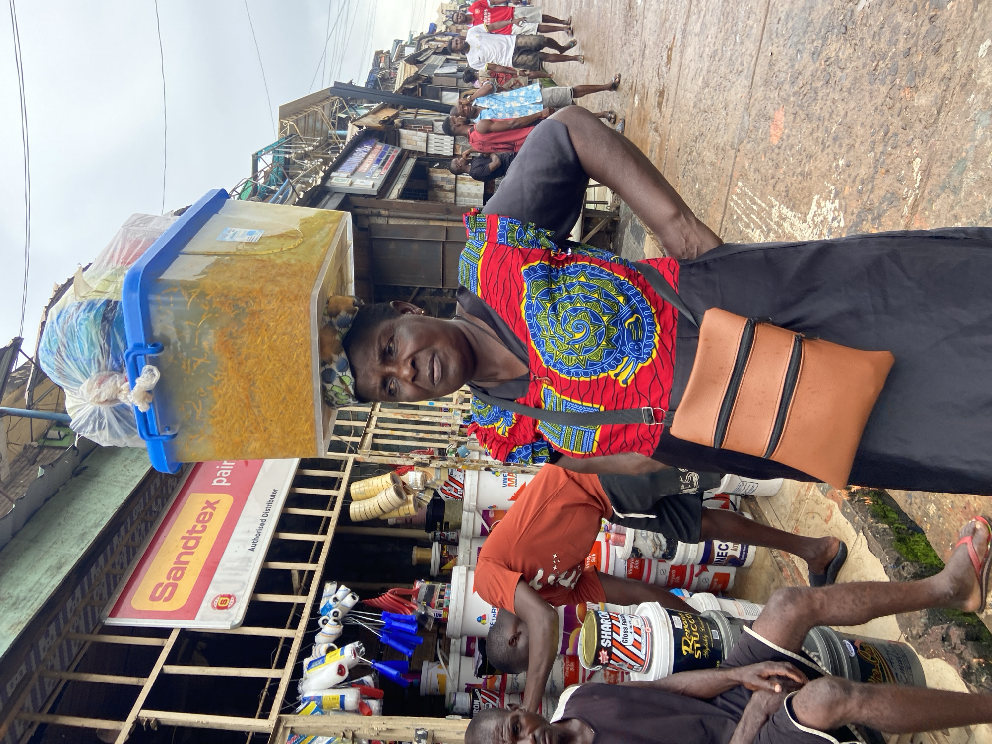 In Ogbunike, Nigeria, a woman with a plastic container filled with yellow items balanced on her head stands in the foreground. She wears a colorful patterned top and a black skirt, with a brown crossbody bag. Behind her, a market scene unfolds with stalls selling various goods, including paint cans and supplies. Several men are visible in the background, some browsing, others walking along the wet pavement. The environment suggests an active outdoor marketplace, possibly during or after a rainfall given the damp ground.