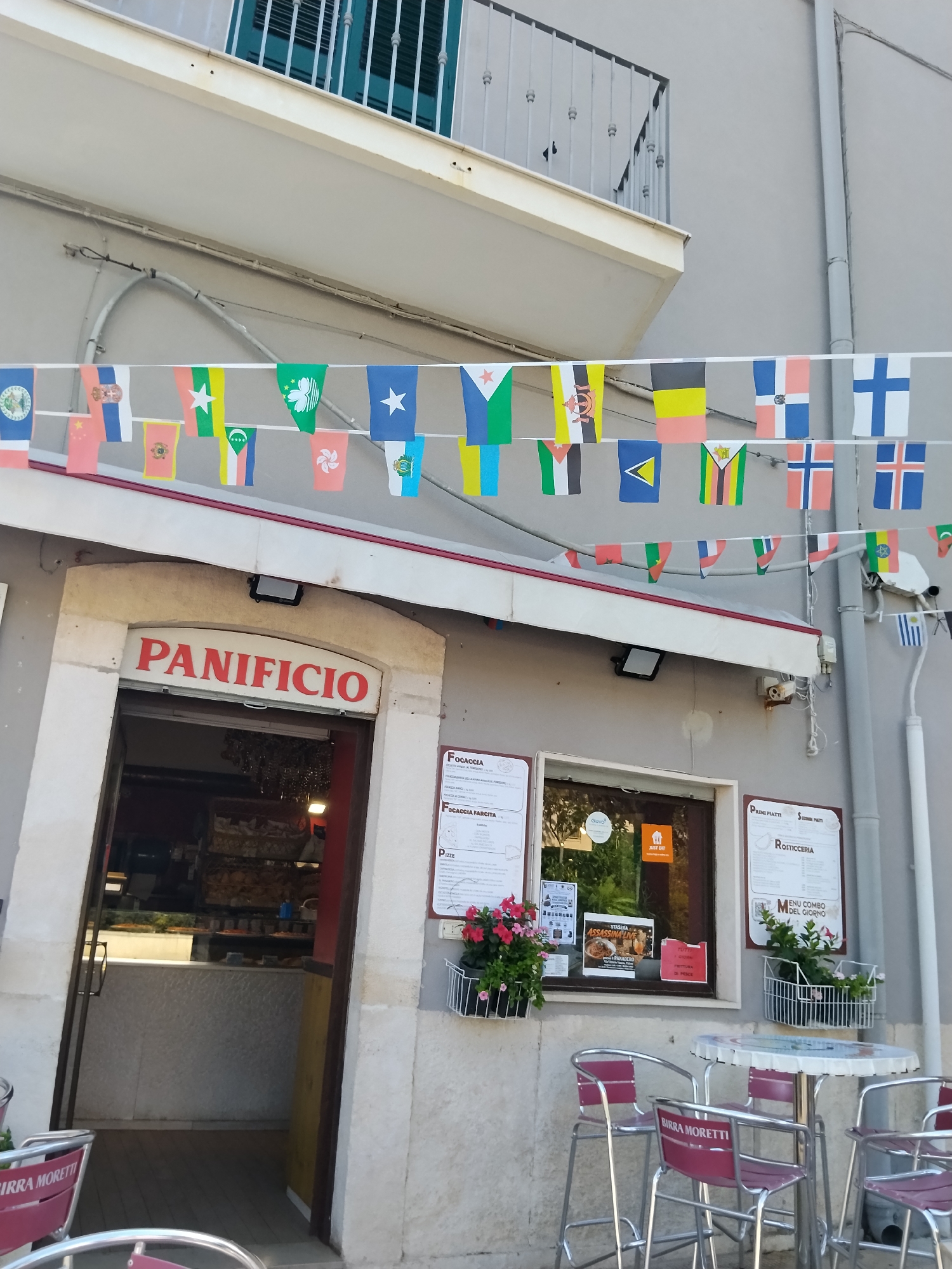 This image shows the exterior of a "Panificio" (bakery) located in Bari, Italy. The bakery has an arched entrance with a red sign displaying the word "PANIFICIO" in white letters. Small flags of various nations are strung across the facade above the entrance. To the right of the entrance, there is a window with menus displayed, including one for "FOCACCIA" and another for "PIZZE." Outside the bakery, there are tables and chairs, suggesting an outdoor seating area. One of the chairs has the "BIRRA MORETTI" logo visible. The building appears to be a few stories high, with a balcony on the upper level. The overall scene depicts a typical street-level establishment in an Italian city.