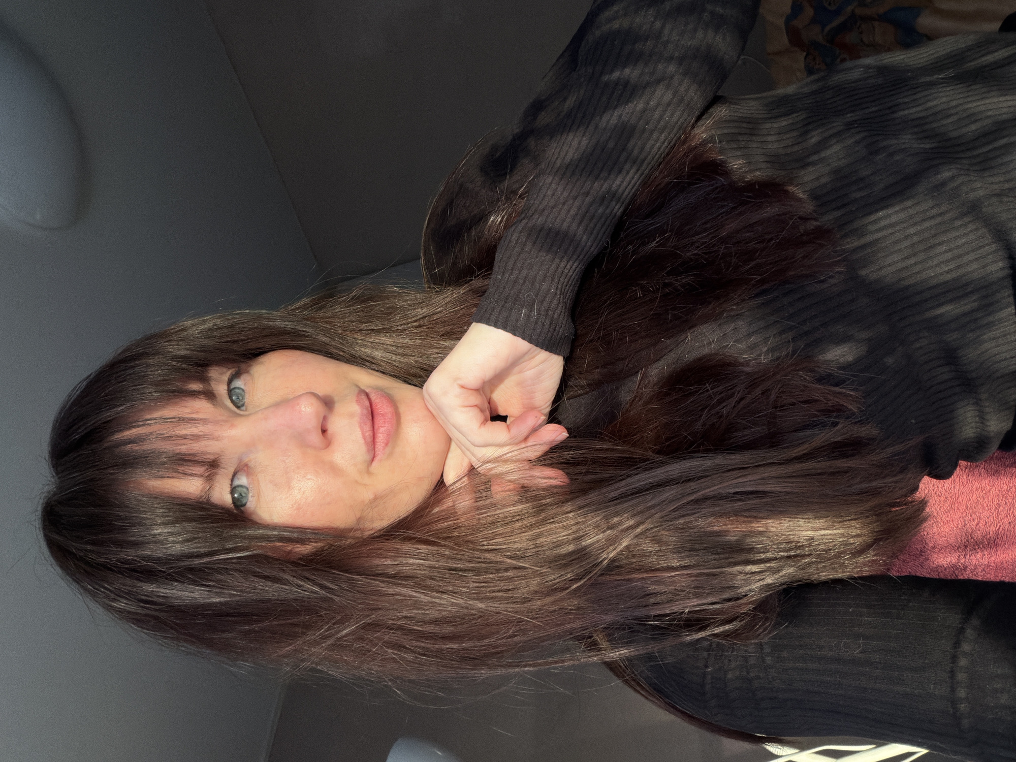 A woman with fair skin, light blue eyes, and dark brown hair with bangs is the subject of the image. She wears a dark, ribbed sweater. Her right hand is positioned with her fingers lightly touching her chin. The background is an interior setting, with a ceiling visible in the top left corner and a wall behind her. A bed with a pink sheet is visible. The location is Perm, Russia.