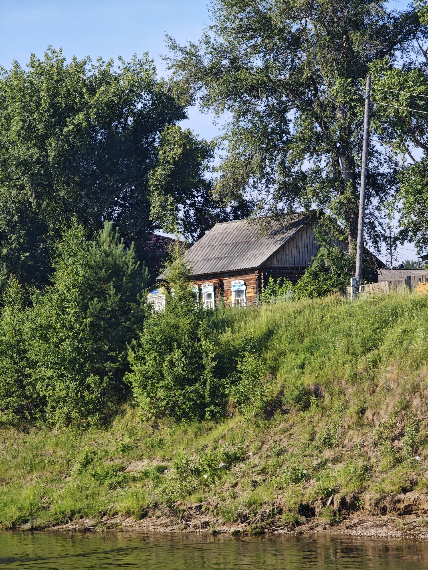 The image depicts a rustic wooden house with decorative window frames situated on a grassy hill overlooking a body of water. Tall trees frame the scene, with a wooden utility pole to the right of the house. The sky is clear and blue. The overall impression is of a tranquil, rural setting, likely in the village of Galanino, Russia, given the architectural style and natural surroundings. There are no people or animals visible in the image.