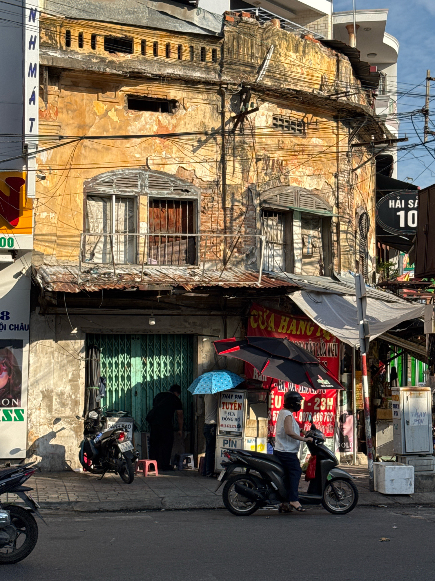 The image depicts a street scene in Nha Trang, Vietnam. In the foreground, a paved road is visible with several motorcycles parked and in motion. A dark-colored motorcycle with a rider wearing a white top and dark pants is prominently featured, facing right. Another motorcycle is parked to the left.
The background showcases an old, weathered building with peeling yellow paint and visible brickwork. The architecture suggests a long history, with ornate window frames and a corrugated metal roof extension. Electrical wires are strung haphazardly across the facade. Green metal gates are closed on the ground floor of the building, and above them, windows with metal bars are visible.
Several signs with Vietnamese text are displayed on and around the building. One sign, red with white lettering, advertises a business. Another sign with the number "10" on a dark oval background is also visible, suggesting a street number or a business identifier. Umbrellas, one blue with white polka dots and another black and red, are set up near the building entrance, possibly for shade or a market stall.
A person in dark clothing is standing near the green gates. Another person, partially obscured by a motorcycle, is also present. A trash bin or storage unit is located on the sidewalk to the right of the building. The overall impression is a bustling urban environment with a blend of old and new structures, typical of many Vietnamese cities.